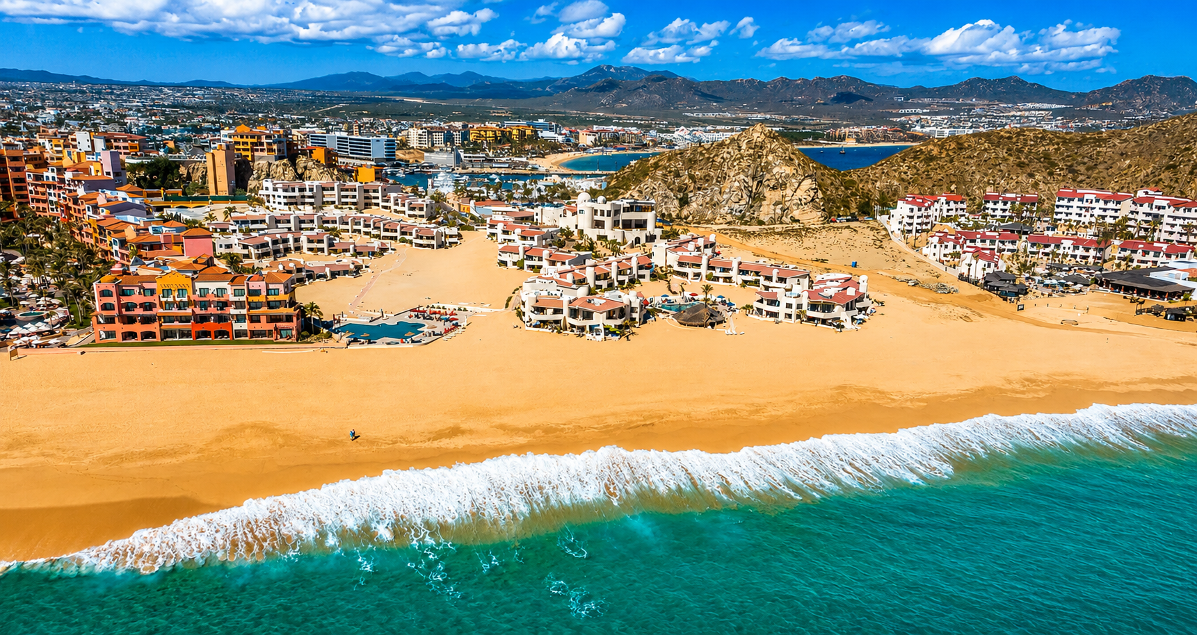 Aerial view of a beachside resort with colorful buildings, sandy beach, ocean waves, mountains in the background, and a cityscape beyond.