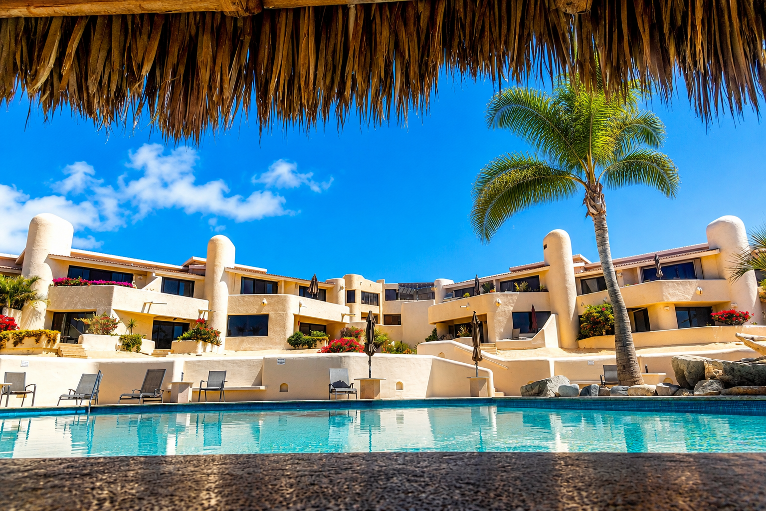 View of a swimming pool at a resort with beige buildings, colorful plants, a palm tree, and clear blue sky