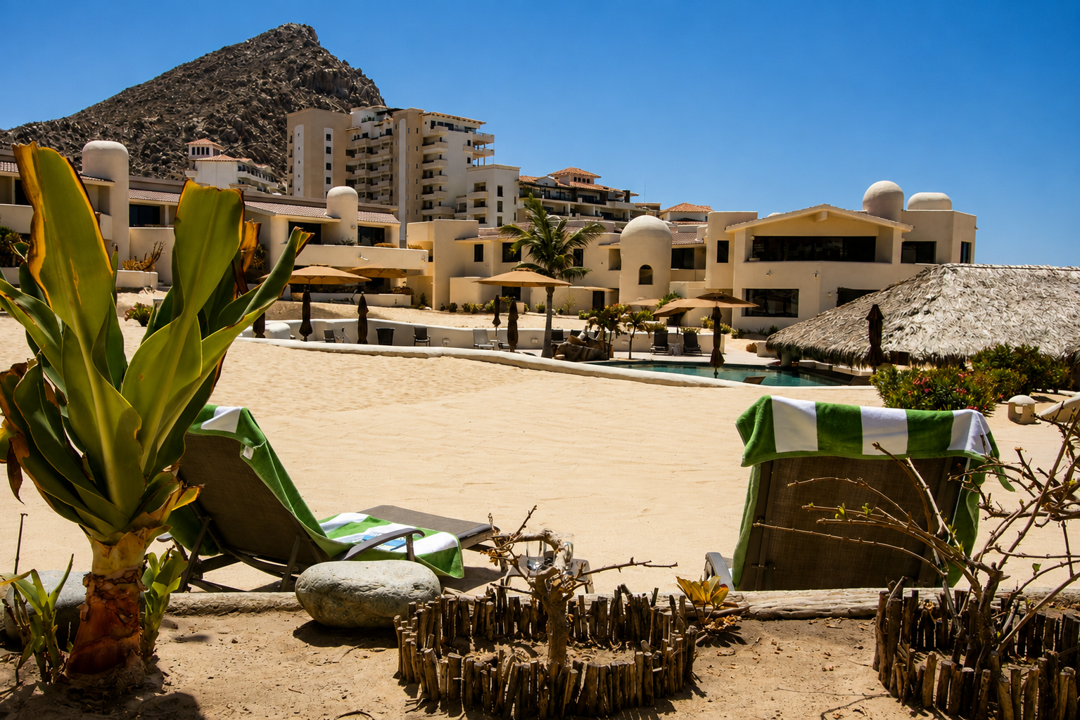 Beachside view with lounge chairs, green and white towels, and tropical plants in the foreground, with resort buildings and a mountain in the background.