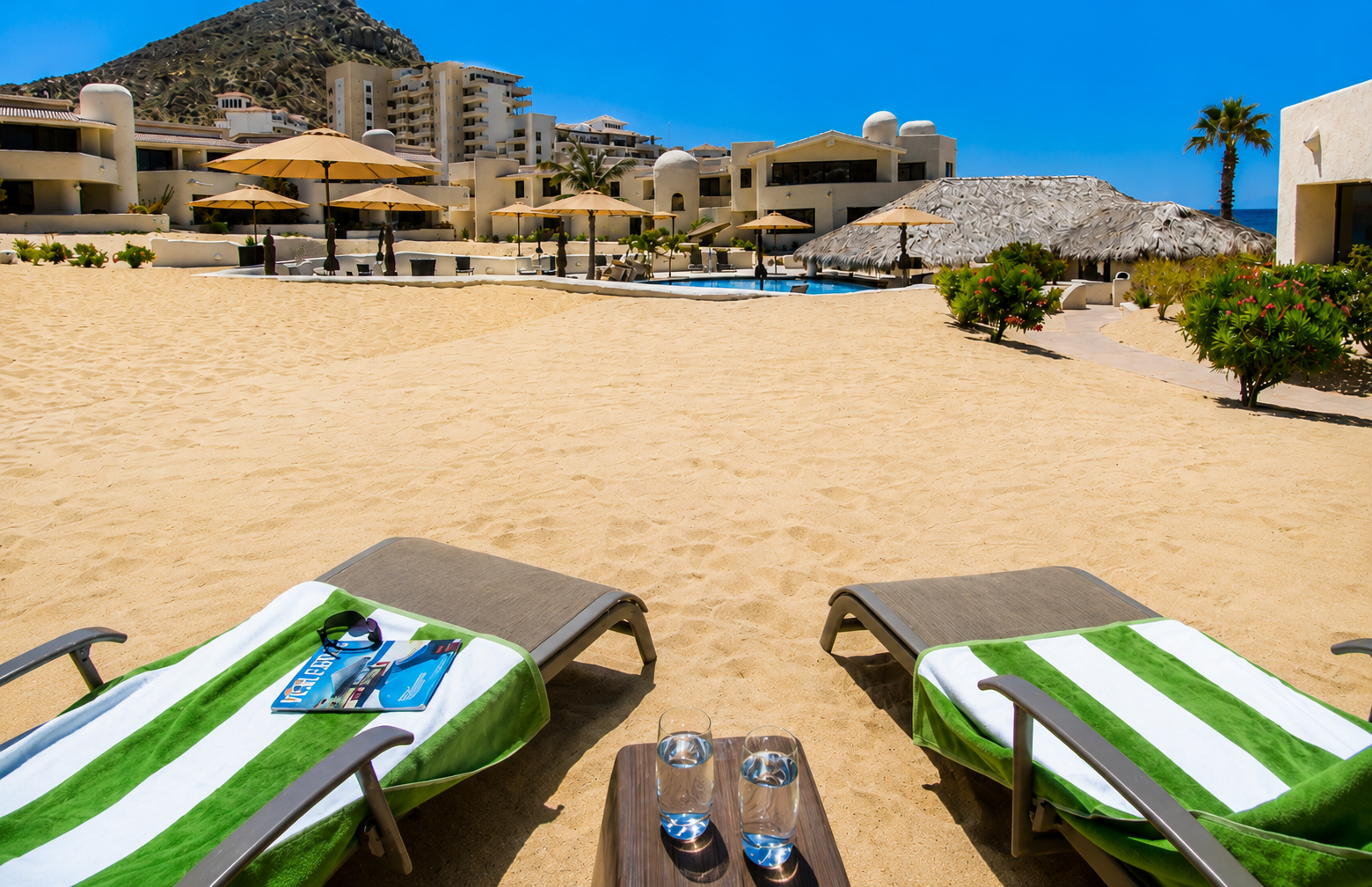 Two lounge chairs with green and white striped towels on a sandy beach, with a small table and two glasses of water in the foreground. In the background, there are umbrellas, a pool, tropical plants, and white buildings with a mountain behind.
