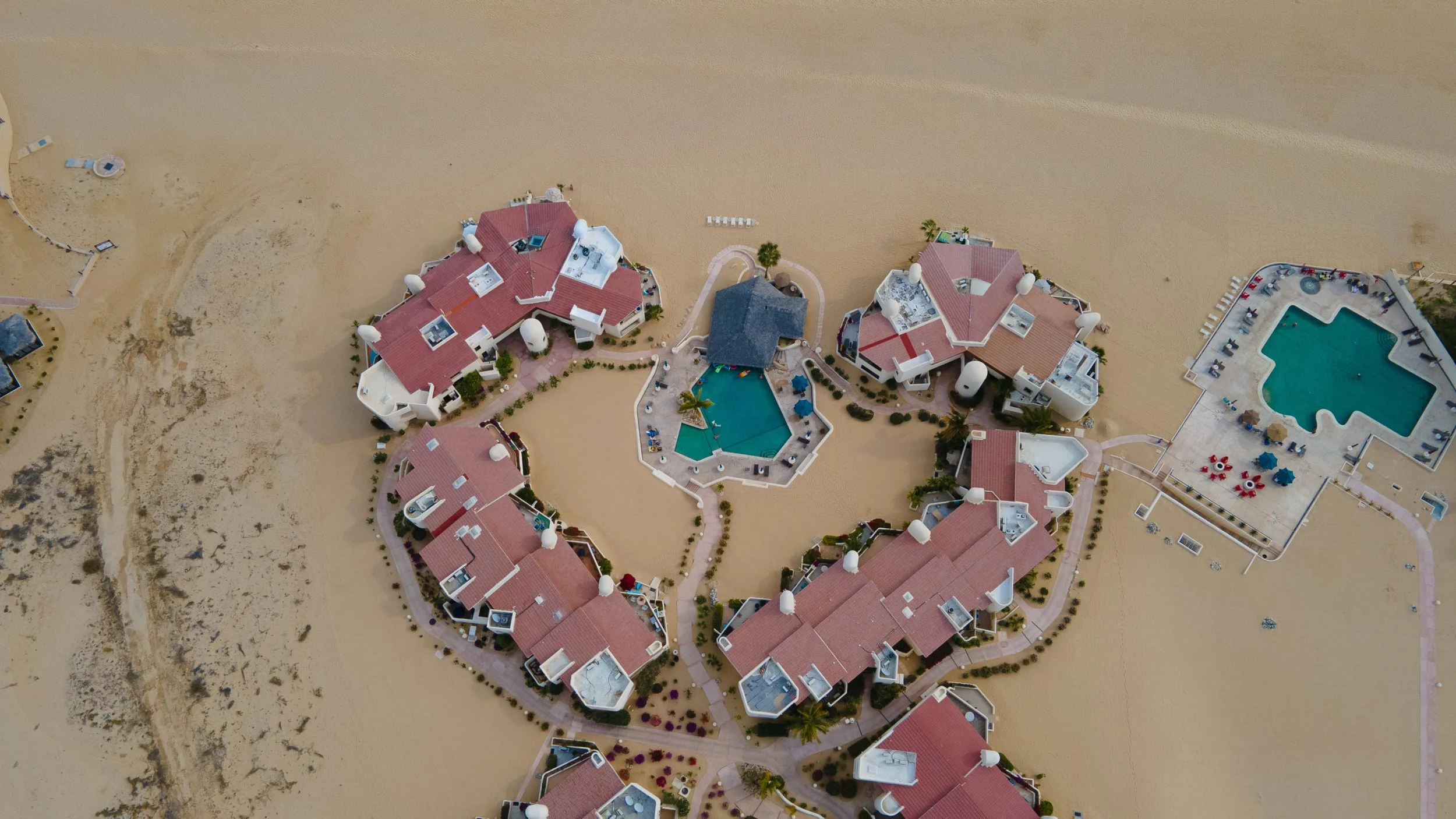 Aerial view of a beachside resort with multiple buildings, swimming pools, and pathways surrounded by sand and some sparse vegetation.