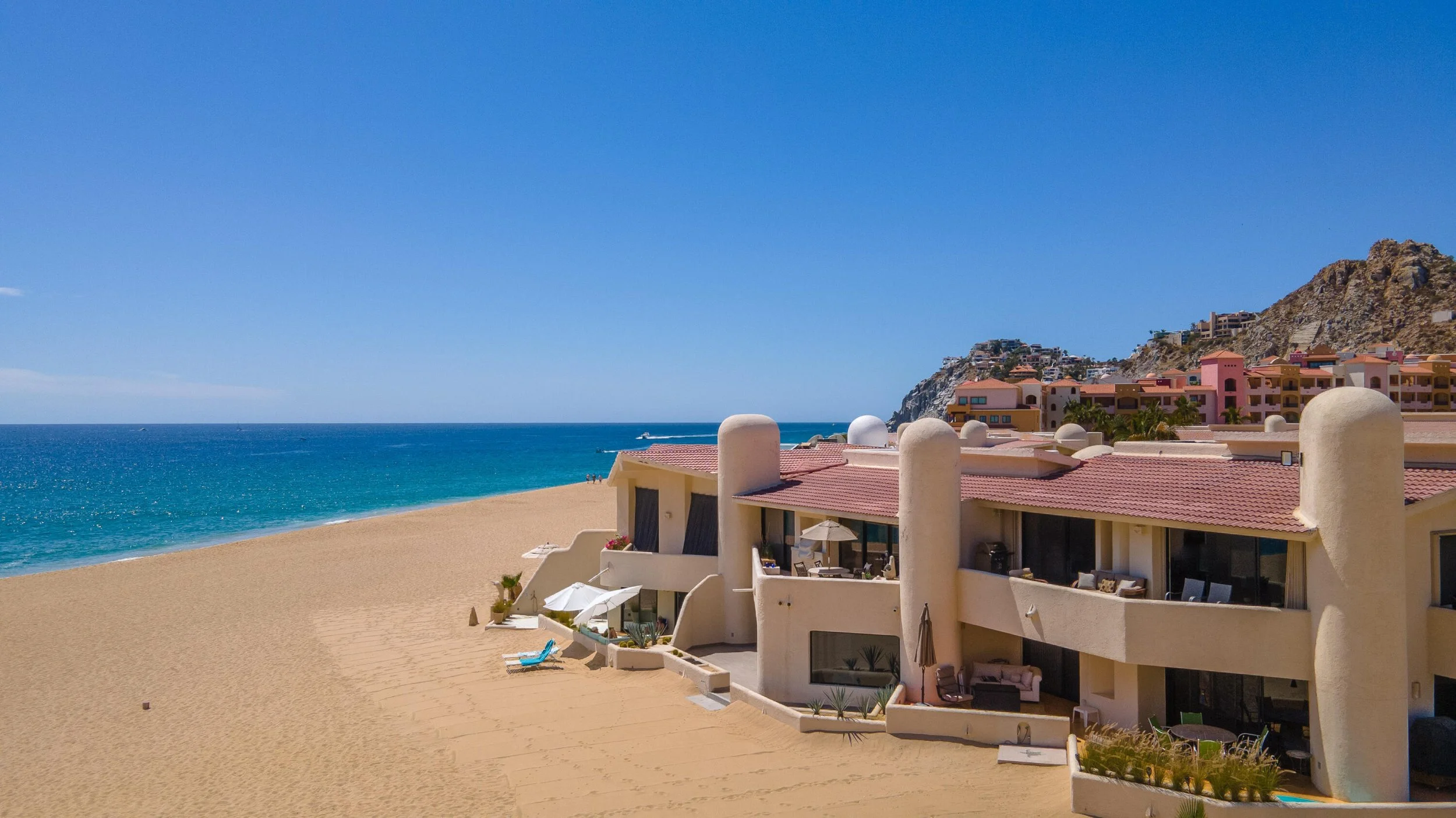 Beachfront resort with white buildings, red-tiled roofs, and outdoor lounging areas, overlooking the sandy beach and blue ocean under a clear sky