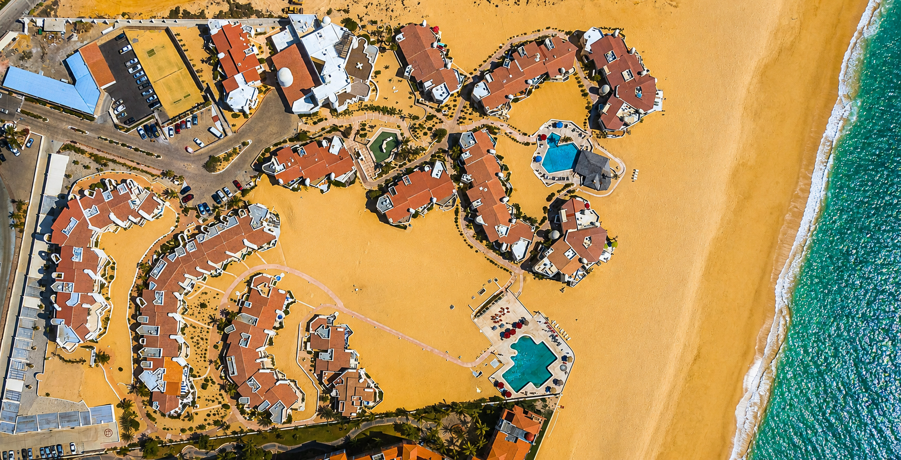 Aerial view of a beachside resort with multiple buildings, pools, parking lot, and sandy beach adjacent to the ocean.