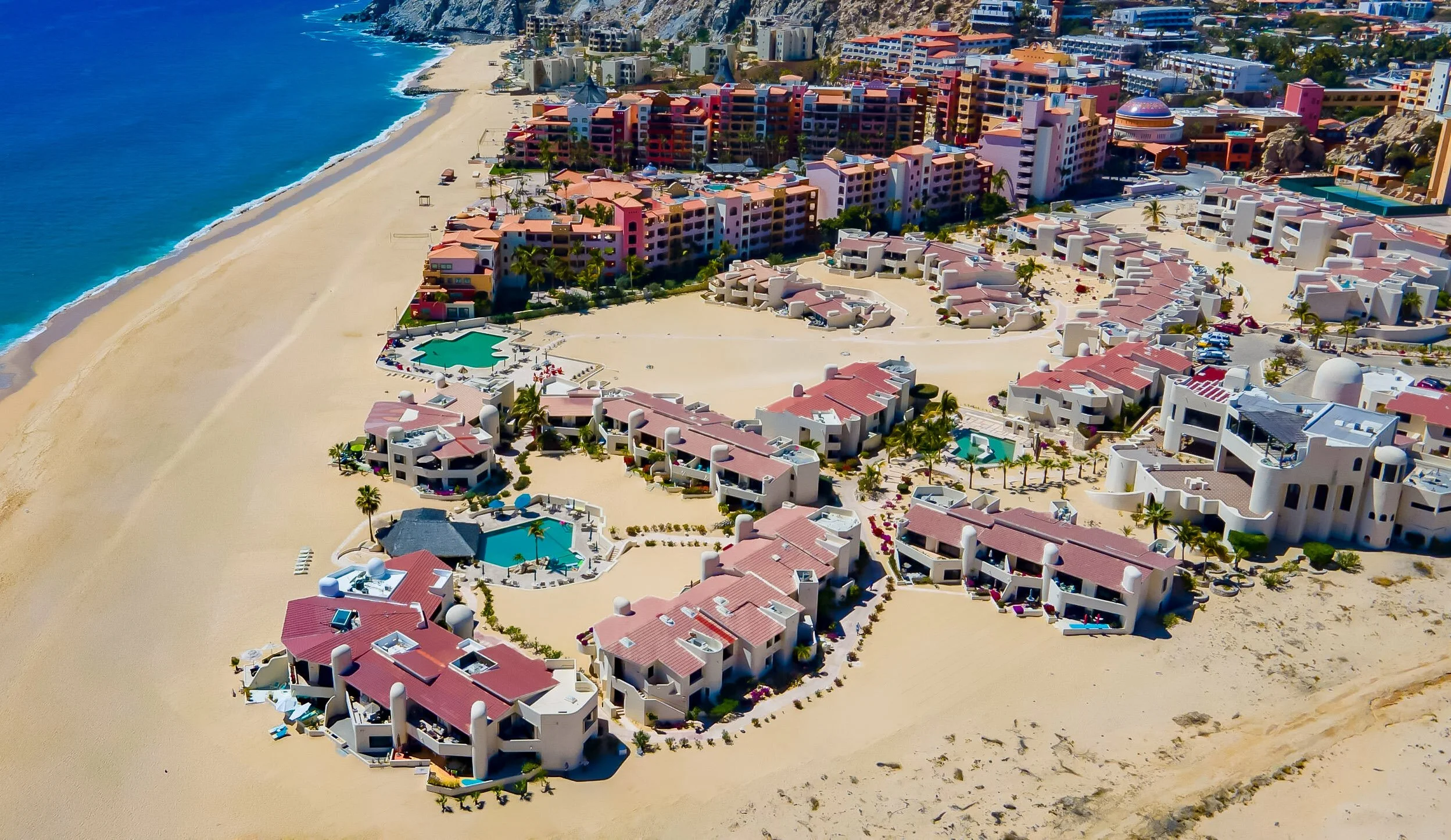 Aerial view of a coastal resort with white buildings, red roofs, and pools along a sandy beach with clear blue ocean.