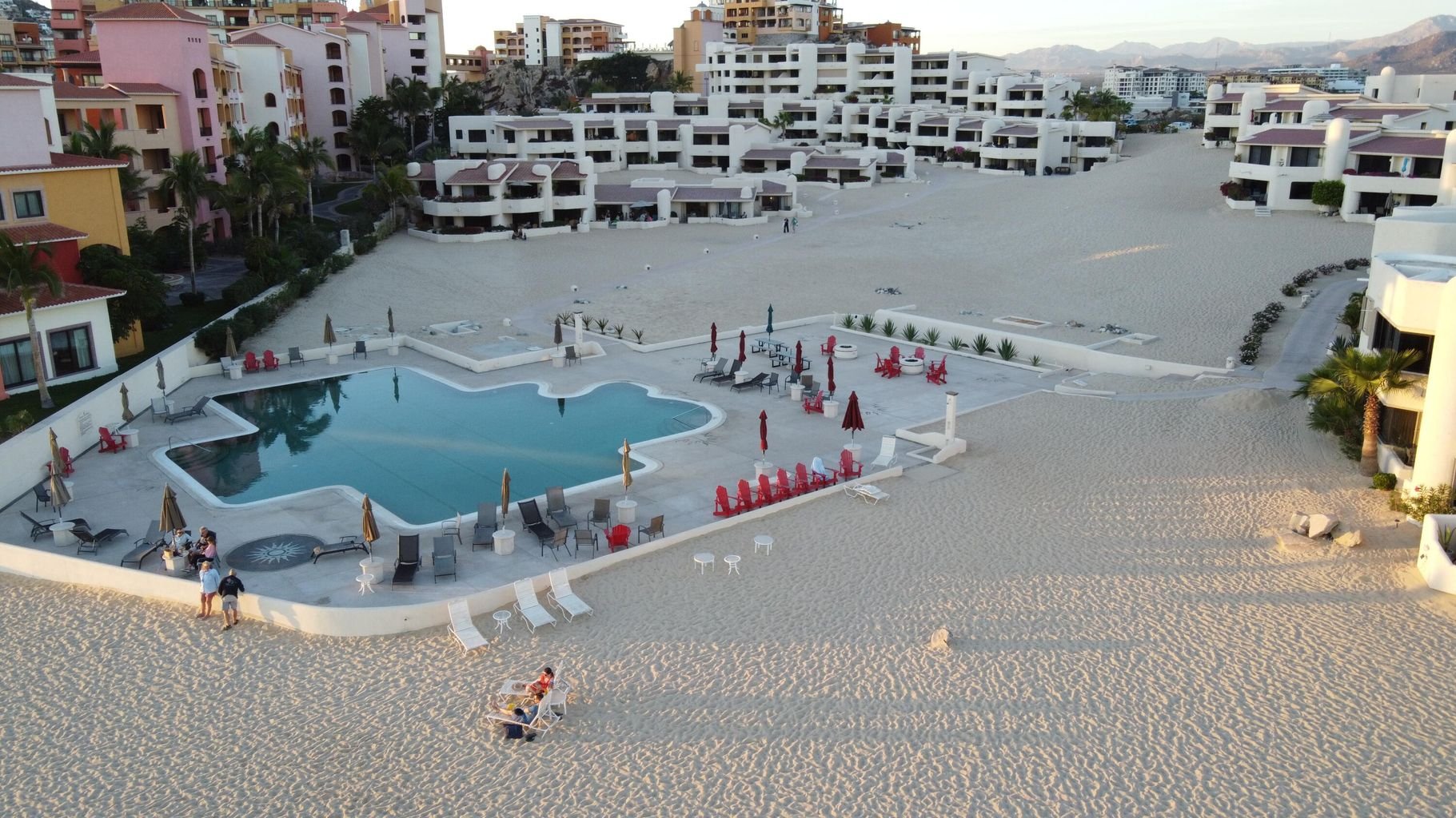 View of a beach resort featuring a swimming pool surrounded by lounge chairs and umbrellas, with multi-story residential buildings and mountains in the background, during late afternoon or early evening.