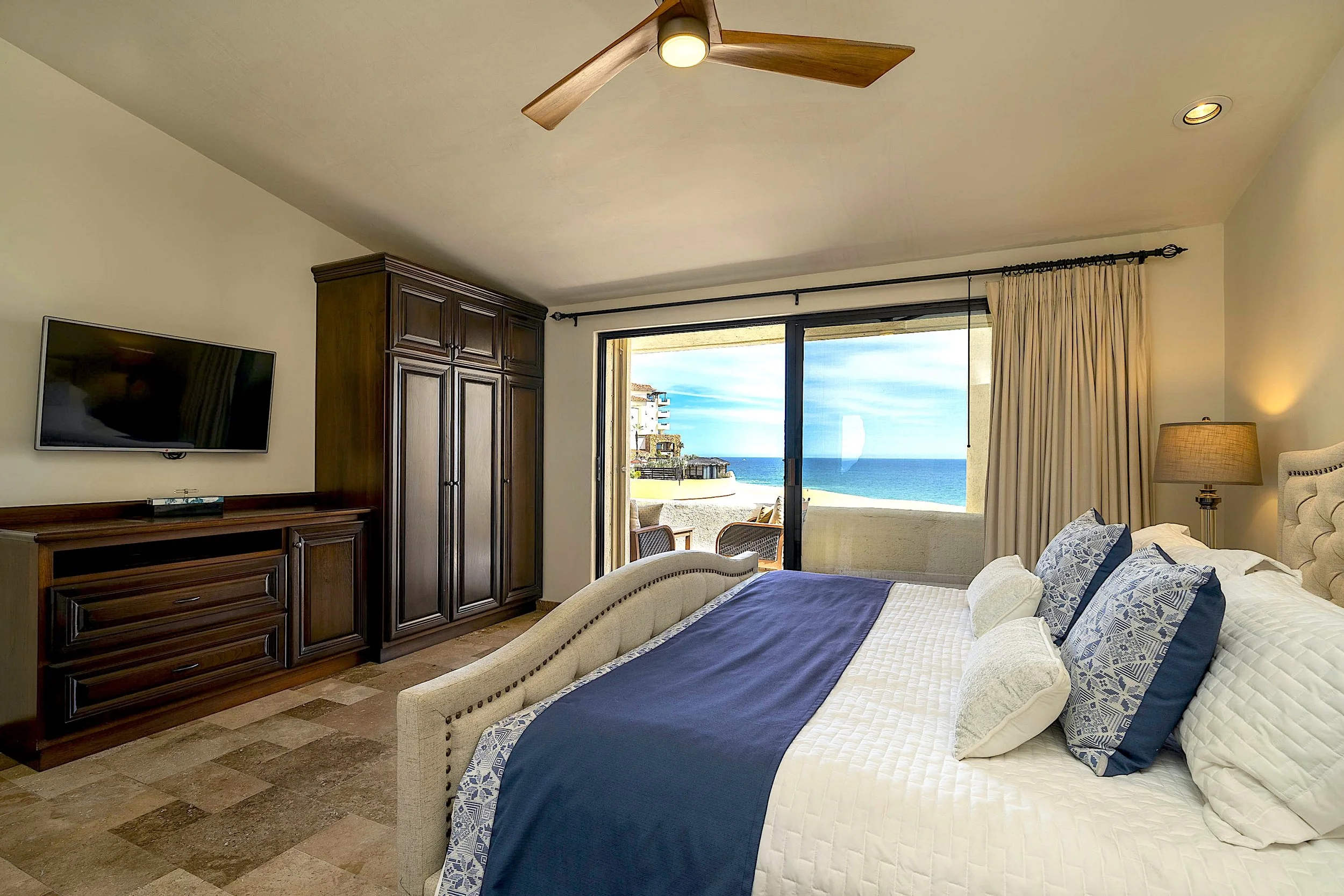 A bedroom with a view of the ocean through sliding glass doors. The room has a large bed with blue and white bedding, a wooden wardrobe, a wall-mounted TV, and a ceiling fan. There are cream-colored curtains and a small outdoor balcony with chairs.