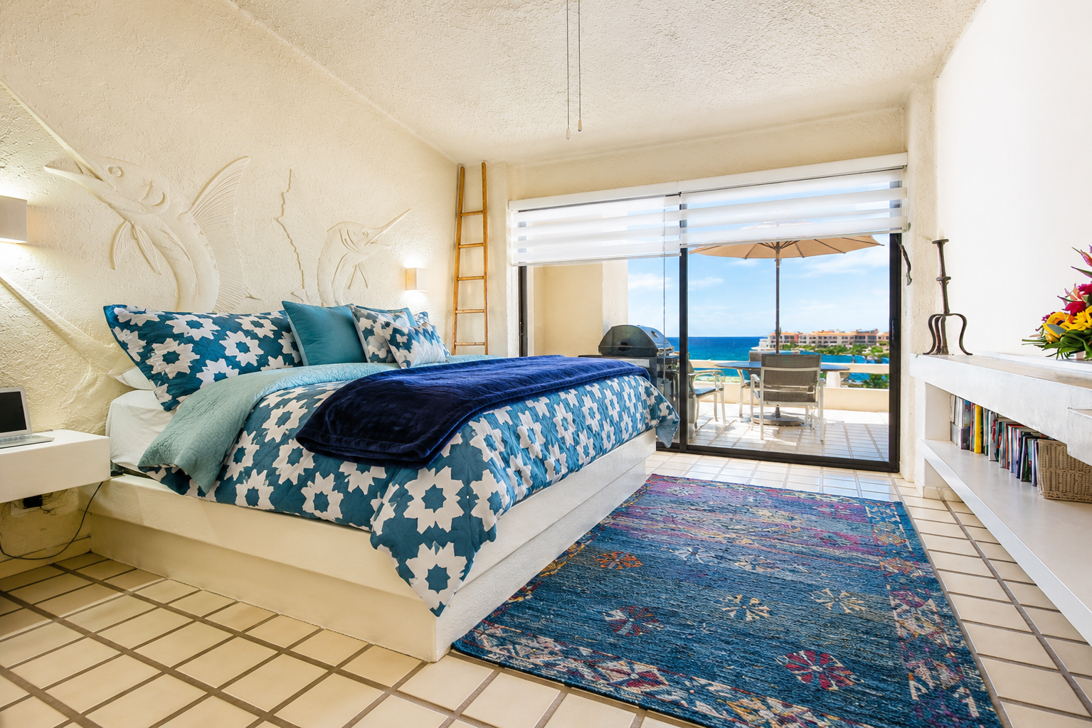 Hotel room with a bed, blue and white patterned bedding, and ocean view balcony with outdoor seating.
