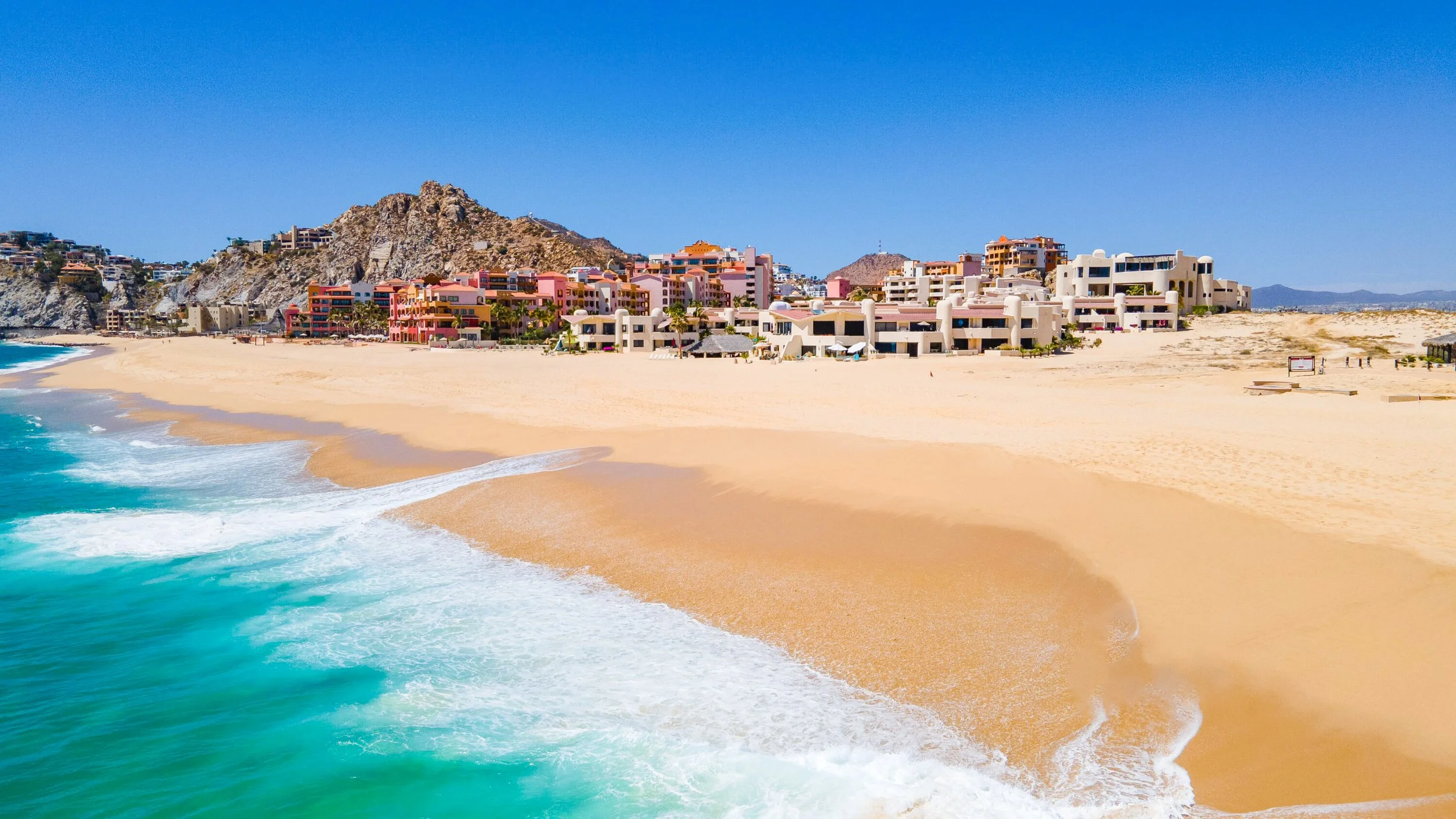 Beach with golden sand, blue ocean waves, and colorful buildings against rocky hills under a clear blue sky.