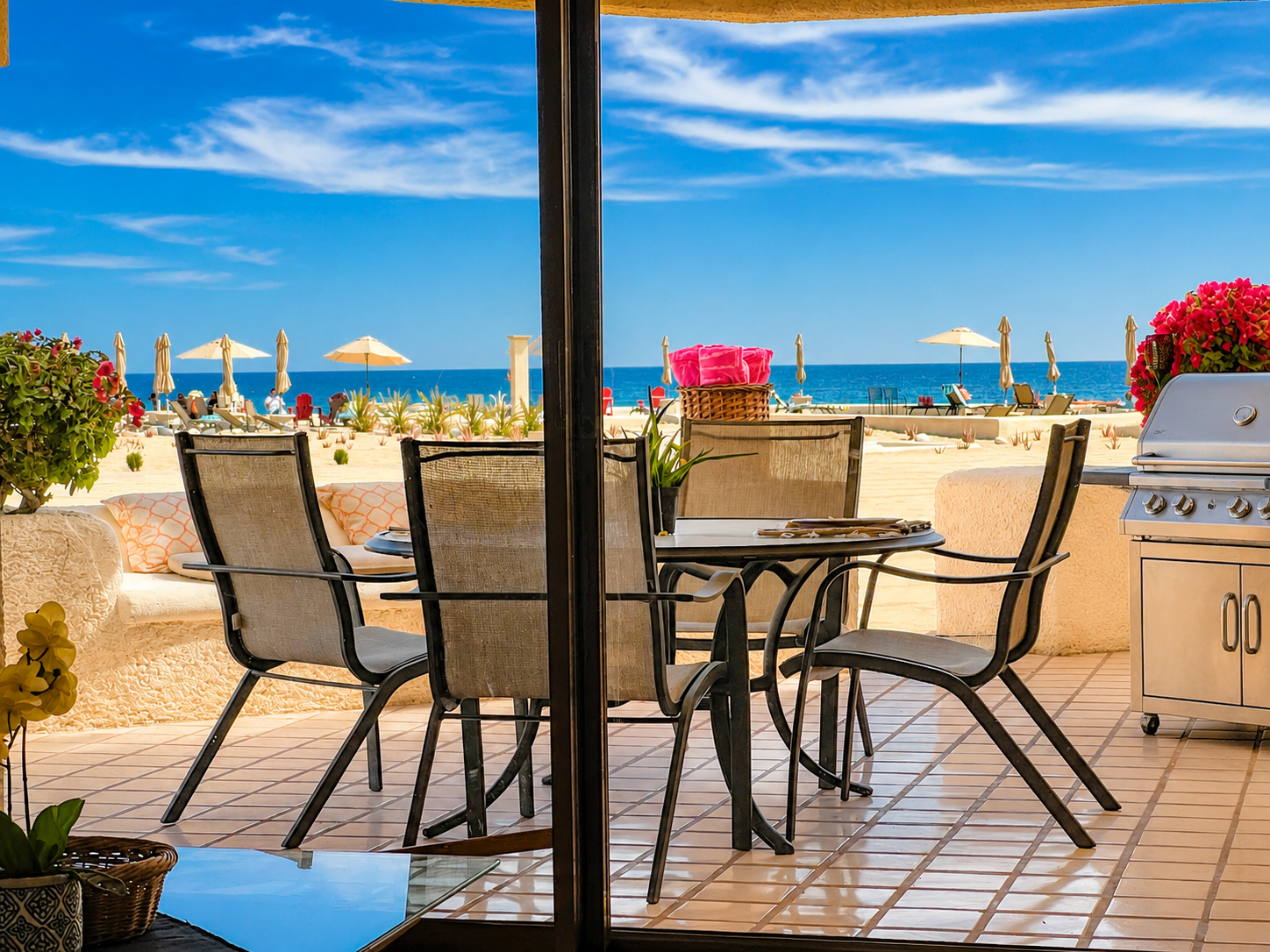 Beach view from a patio with chairs, a table with flowers, and a grill, overlooking sand, umbrellas, and the ocean under a blue sky.