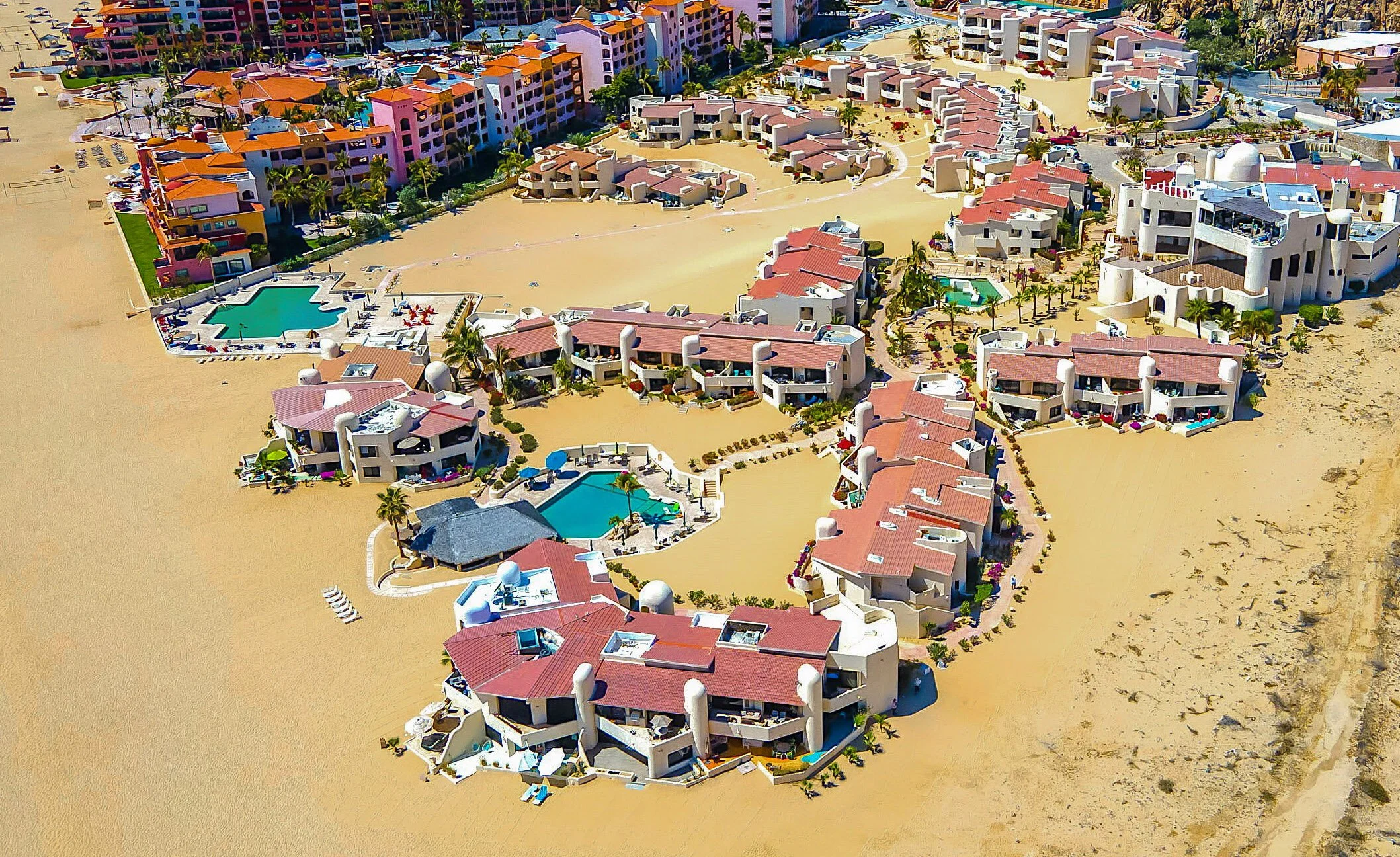 Aerial view of colorful beachfront resort with white buildings, red roofs, swimming pools, sandy areas, and palm trees.