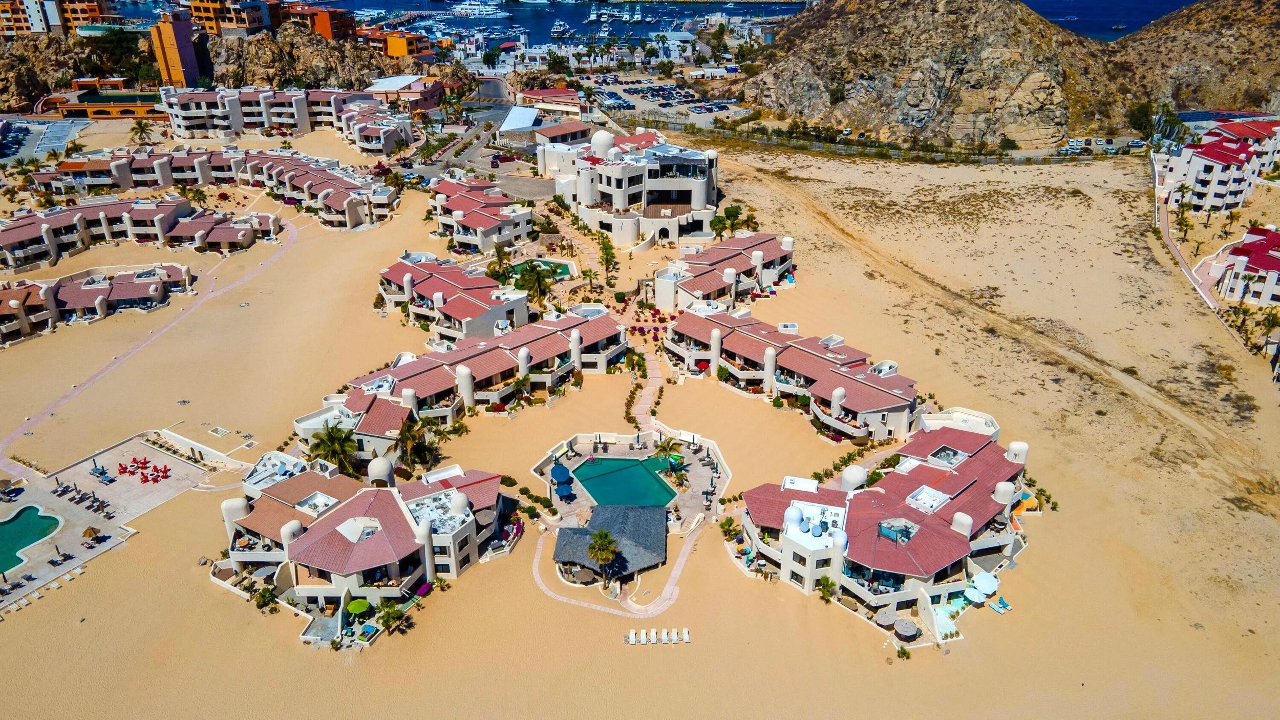 Aerial view of a resort with multiple white buildings and pink roofs, pools, palm trees, and sandy areas near a coastline with boats in the water.