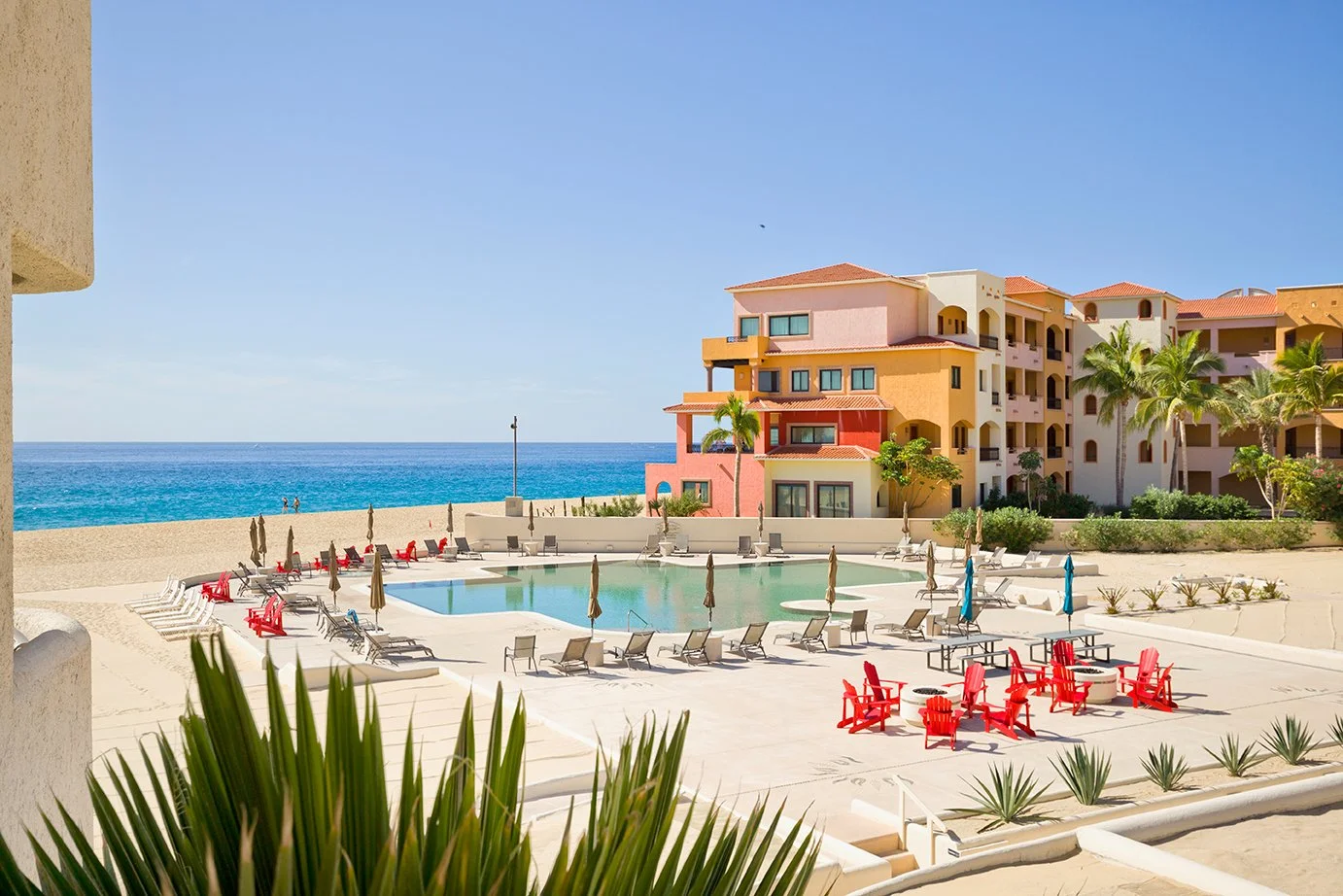 Beachside resort with swimming pool, tropical palm trees, colorful buildings, lounge chairs, and umbrellas under a clear blue sky.