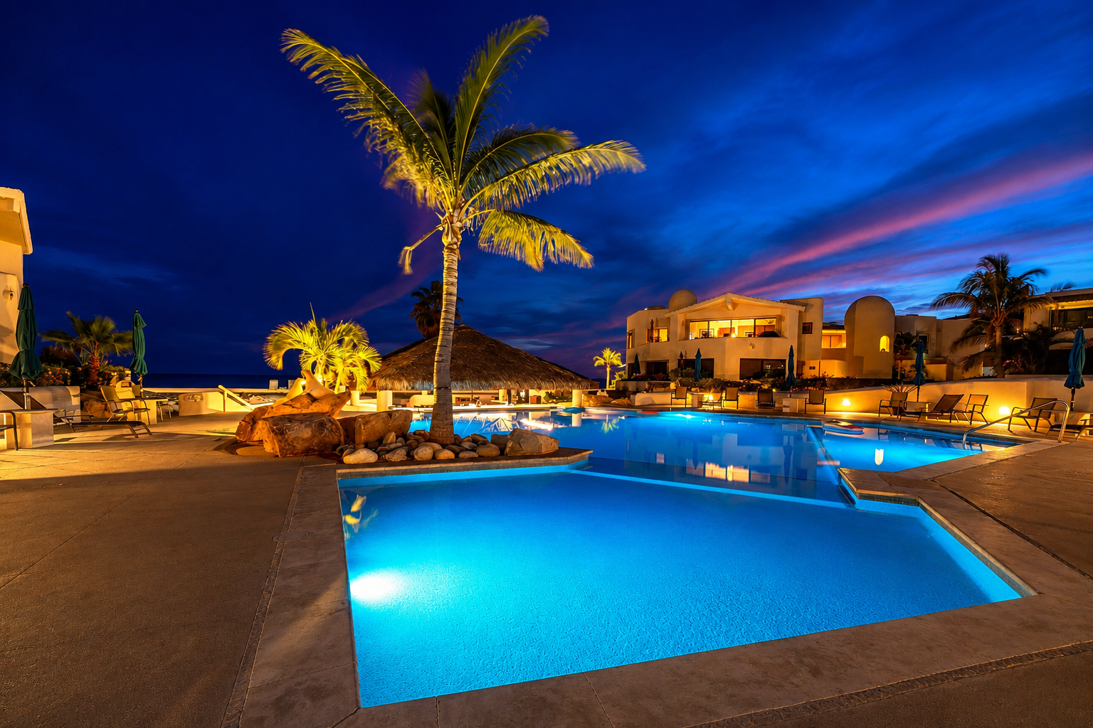 Nighttime scene of a illuminated hotel swimming pool area with palm trees, lounge chairs, a thatched hut, and modern buildings under a colorful cloudy sky.