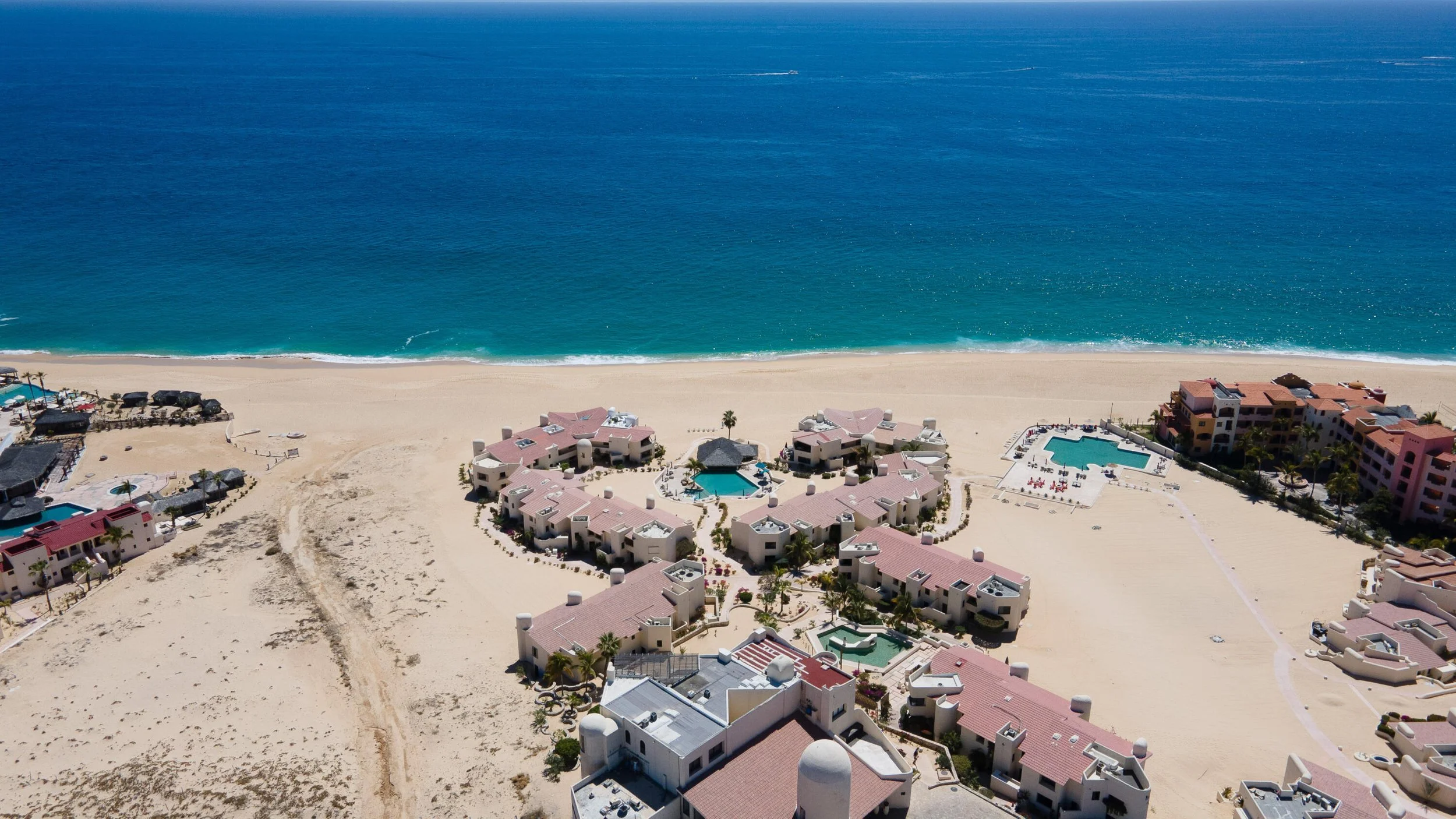 Aerial view of a beachside resort with pink-roofed buildings, swimming pools, and sandy shores next to the ocean.