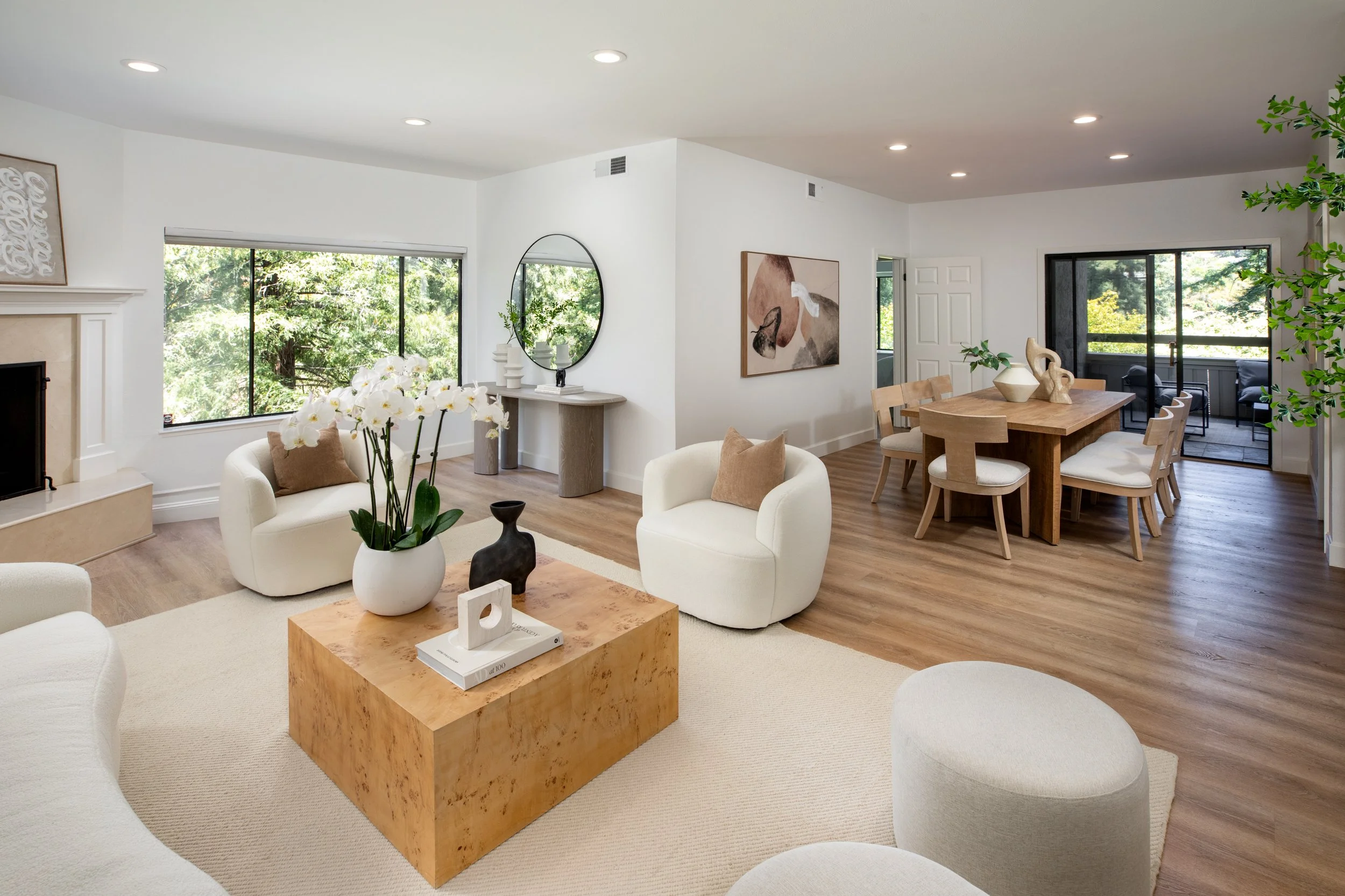 Interior of a living room with white walls, large windows with a view of greenery, and wooden flooring, featuring a seating area with white chairs and coffee table, decorative art, and a dining area with a wooden table and chairs.