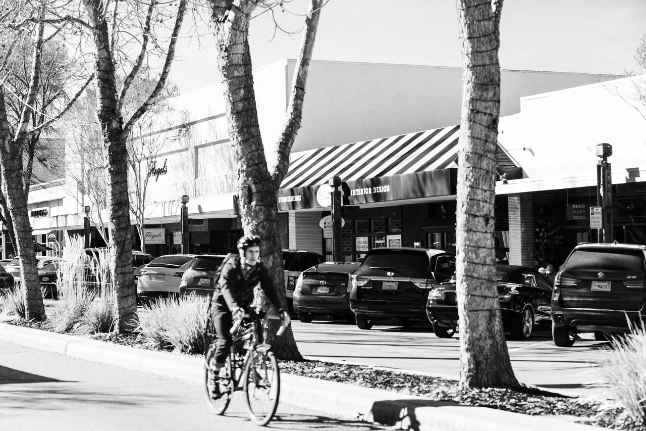 A person riding a bicycle past a parking lot with several parked cars and storefronts, some trees, and an awning in an urban shopping area.