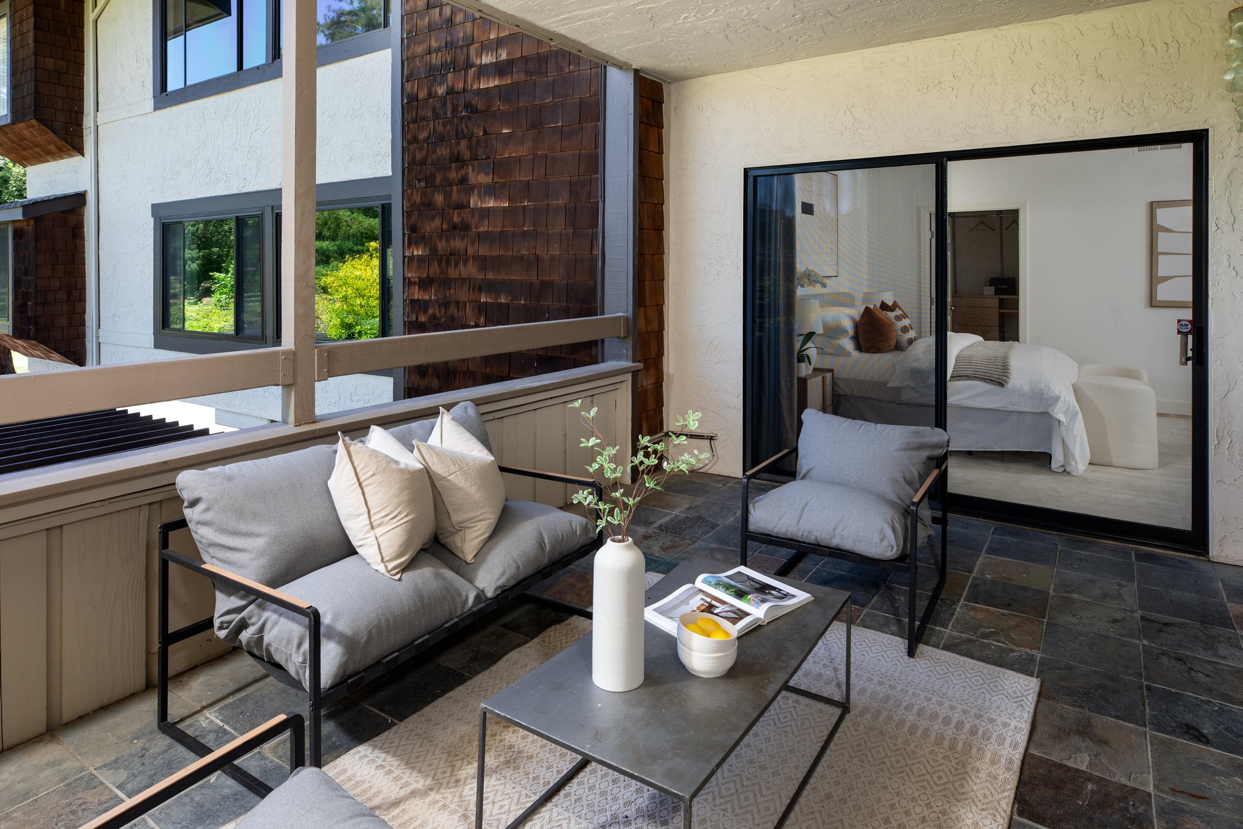 Balcony with outdoor seating area, featuring a sofa with pillows, a coffee table with a book and a bowl of lemons, and potted plants, overlooking neighboring buildings and trees through the railing.