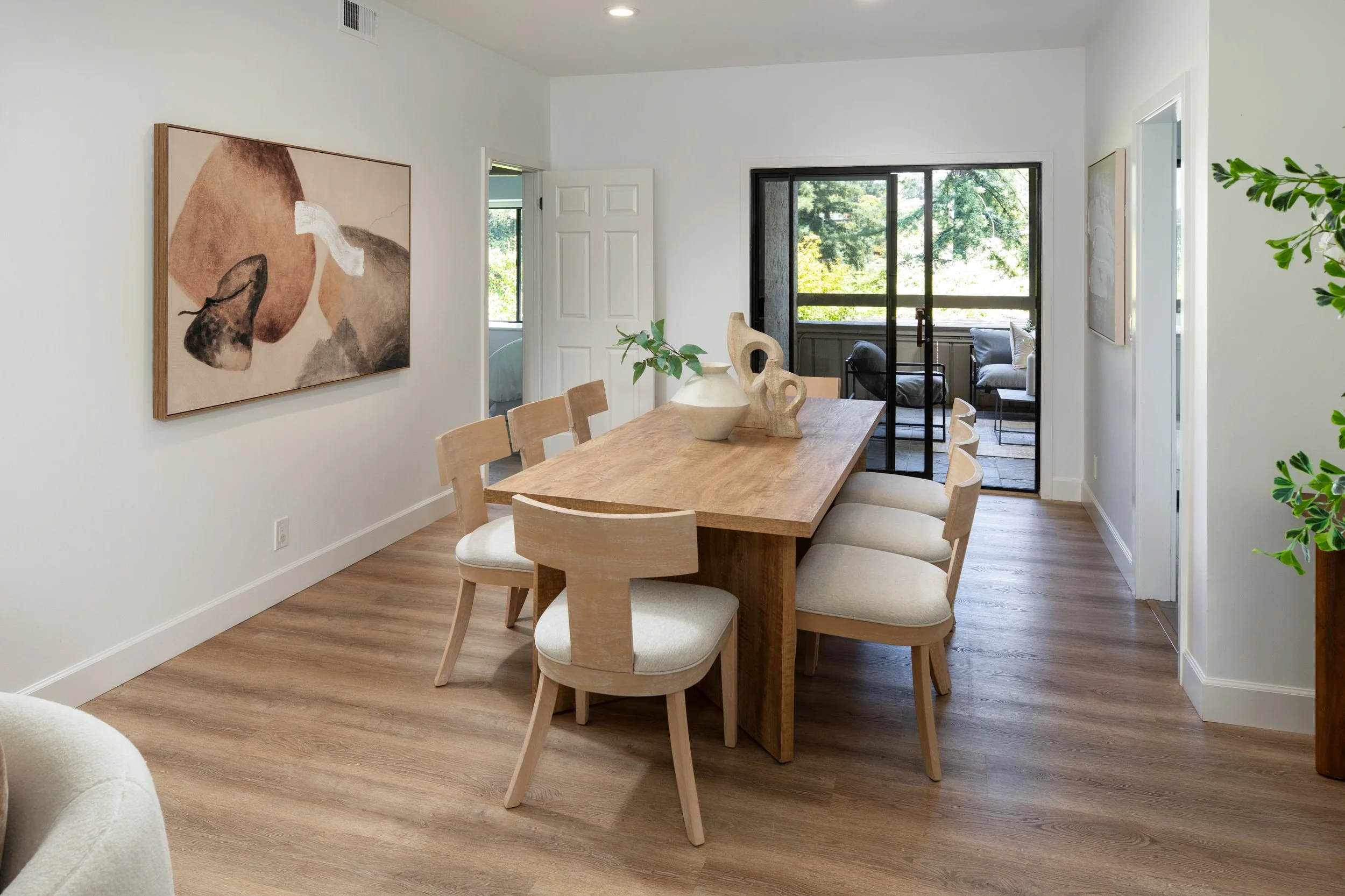 A dining area with a wooden table and six matching chairs with cream cushions, decorated with vases and a small branch plant, adjacent to a living room with seating outside a sliding glass door, with artwork on the white wall and wood flooring.