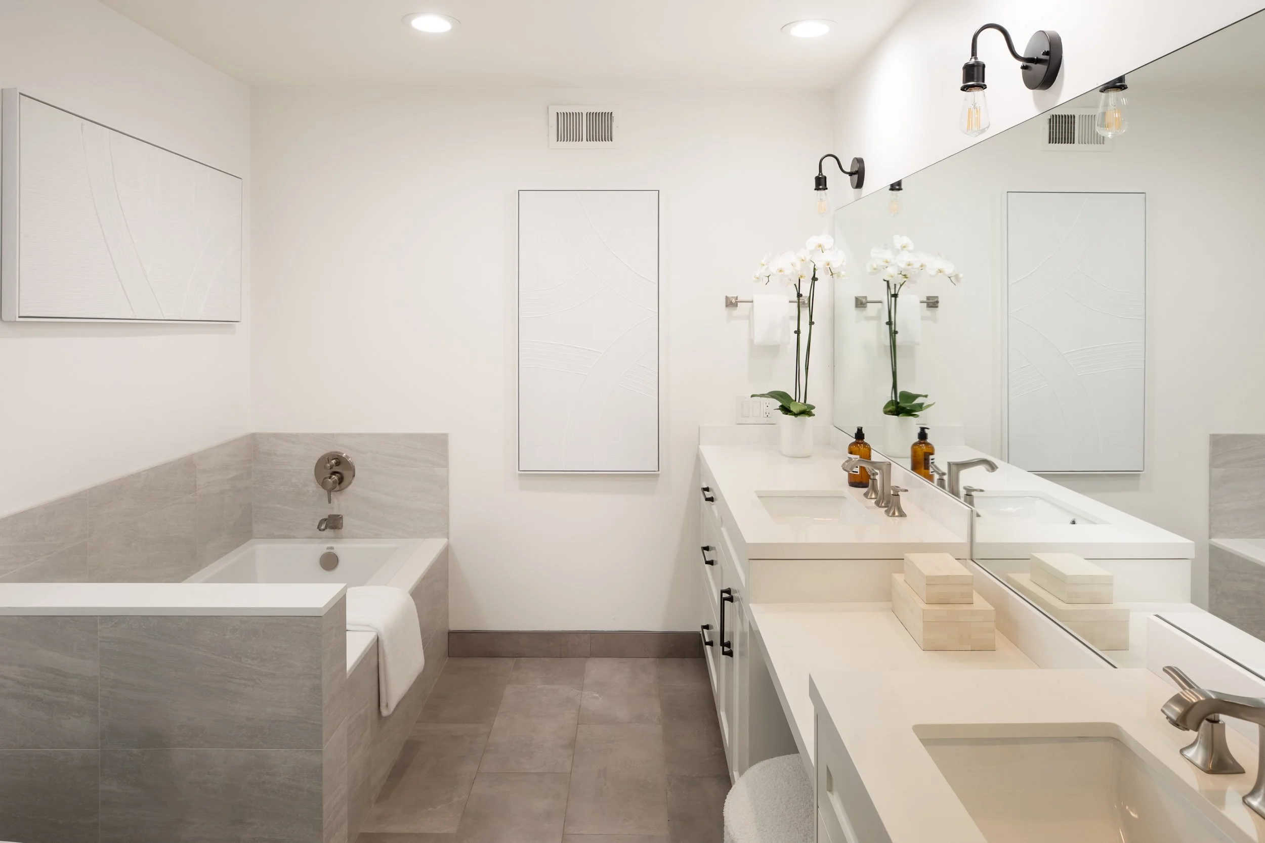 A modern white bathroom with a bathtub, a vanity with a sink, a large mirror, and decorative white orchids.