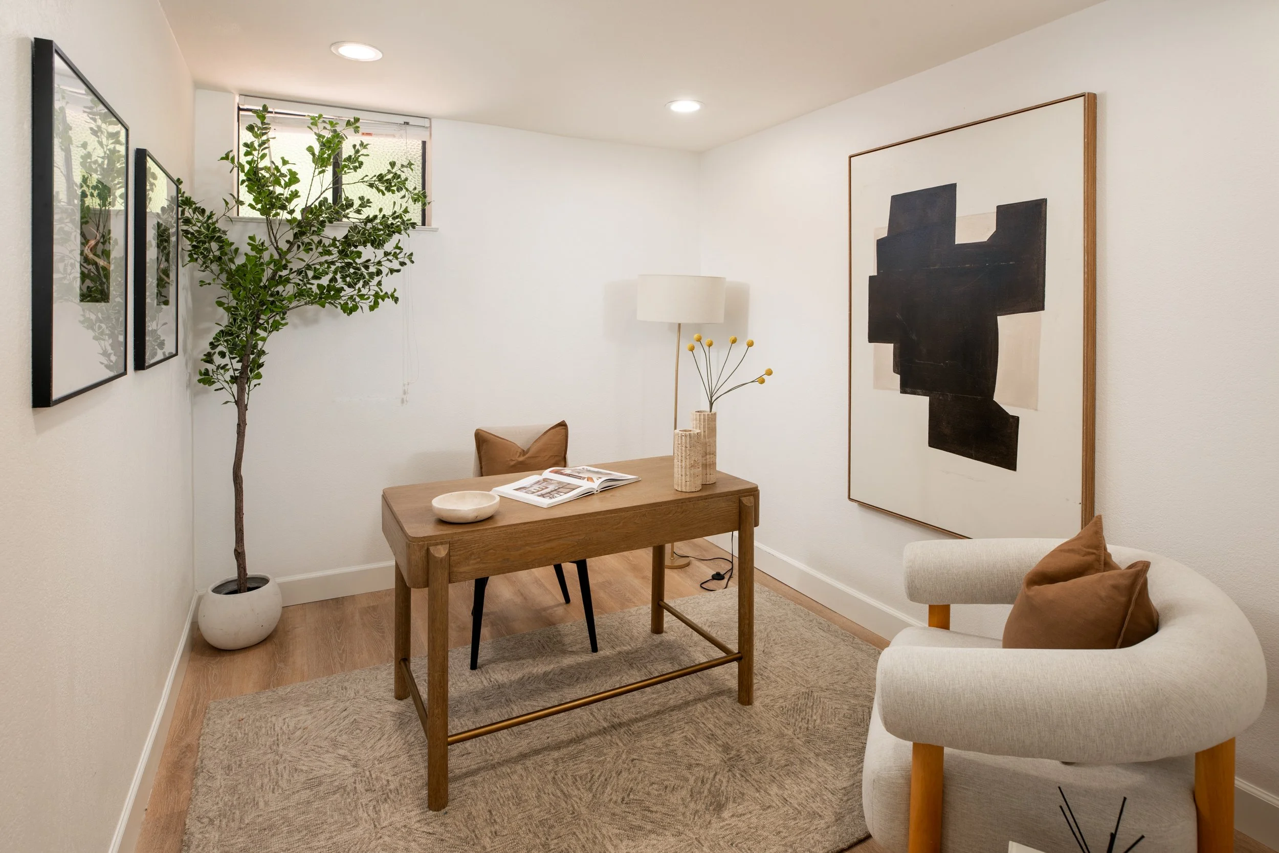 A cozy living room corner with a small wooden table, a white armchair with a brown cushion, a beige area rug, a large potted green plant near a small window, wall art including two framed pictures and a large abstract painting, a tall white floor lam