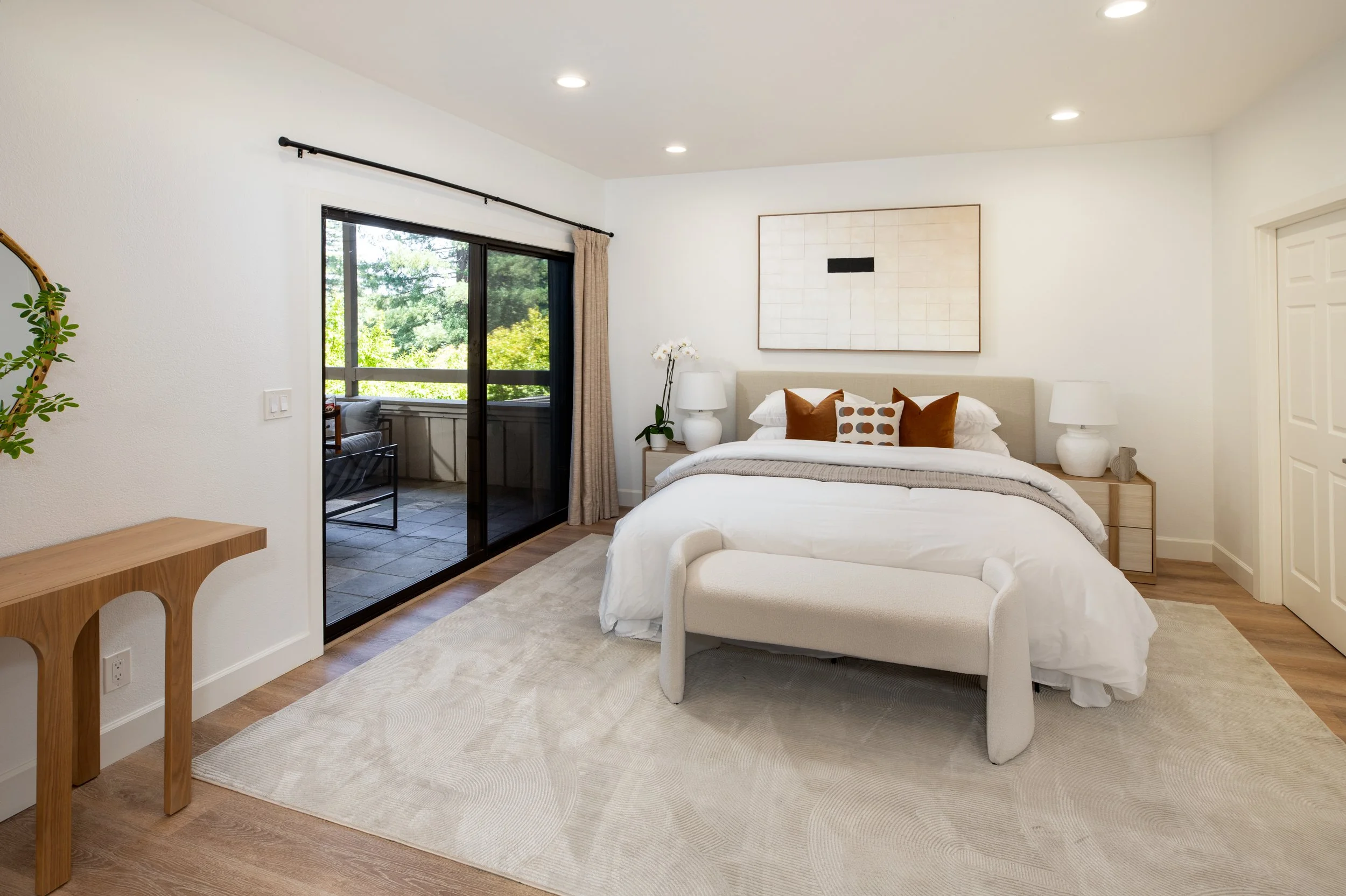Modern bedroom with a large bed, beige headboard, white bedding, and decorative pillows. French doors open to a balcony with greenery outside. Interior includes nightstands, lamps, wall art, and wooden flooring.