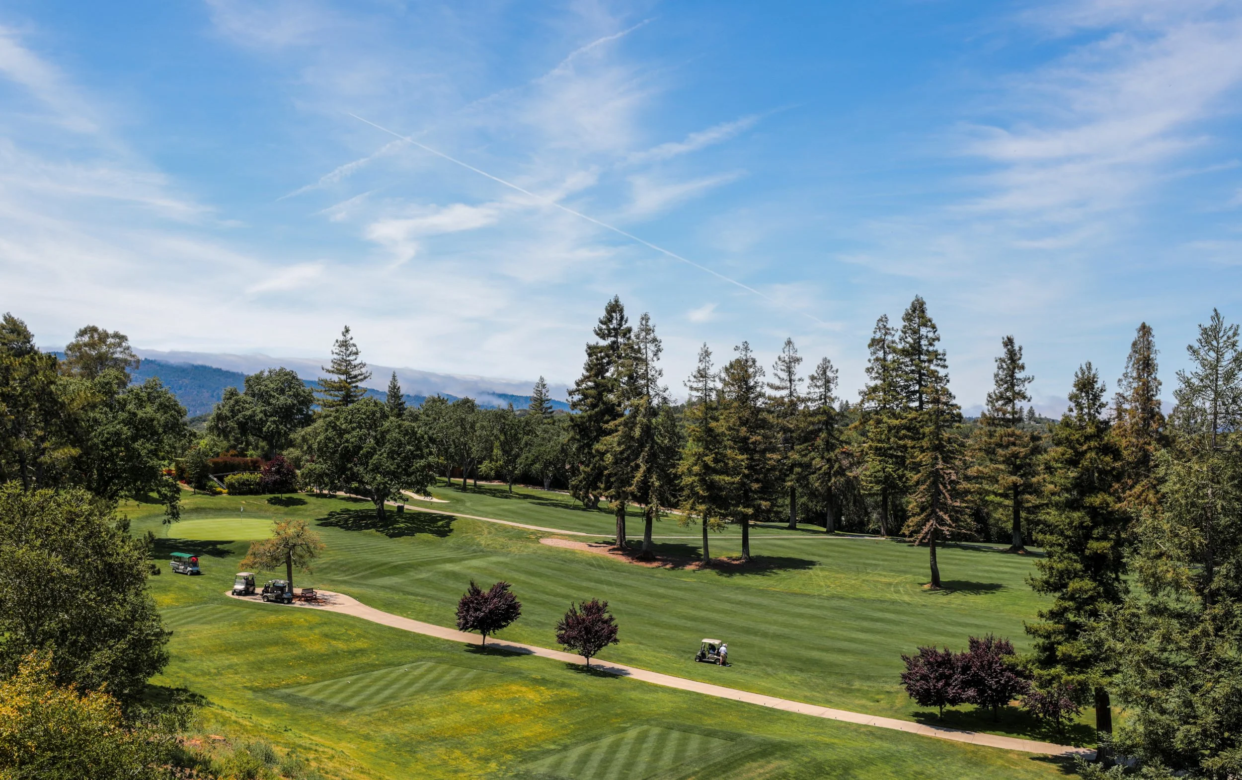 A scenic view of a golf course with green fairways and surrounding trees, under a blue sky with some clouds.