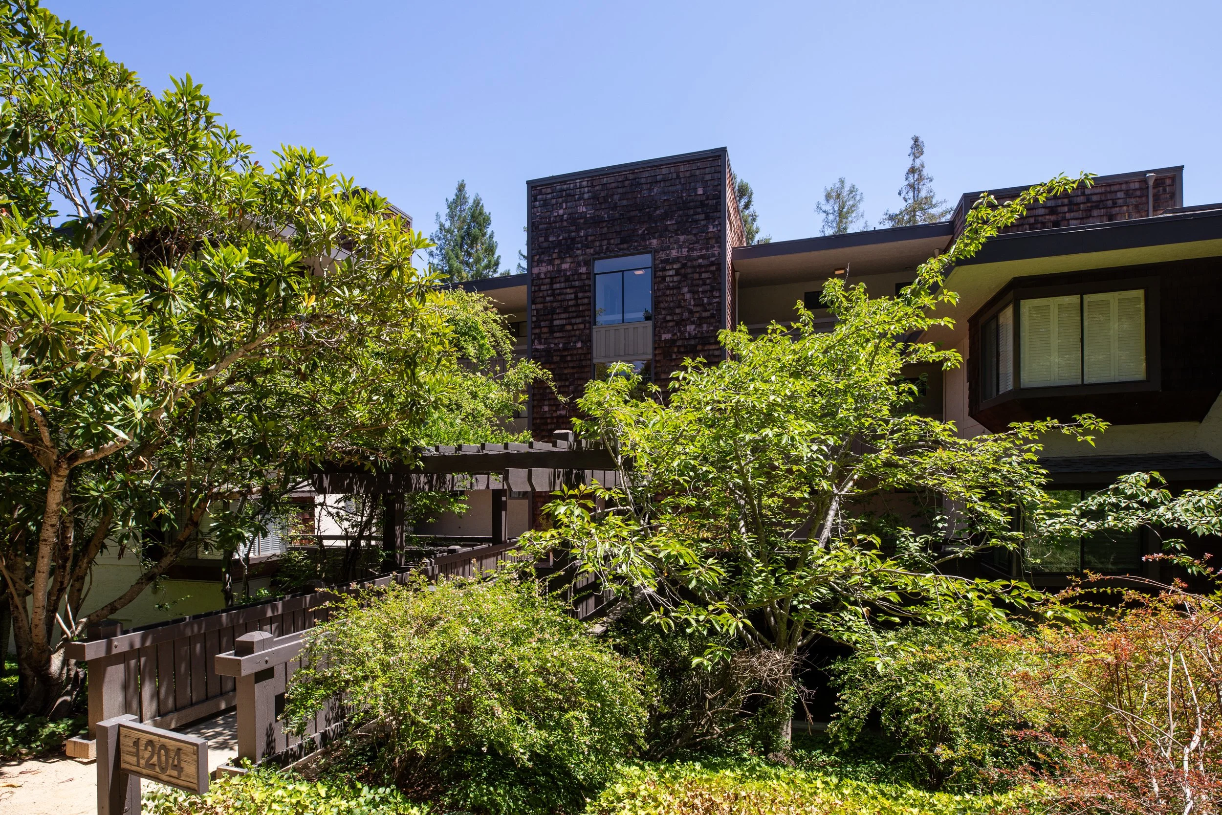 Multi-story residential building with brown wooden siding, large windows, and a balcony, surrounded by green trees and shrubs under a clear blue sky.