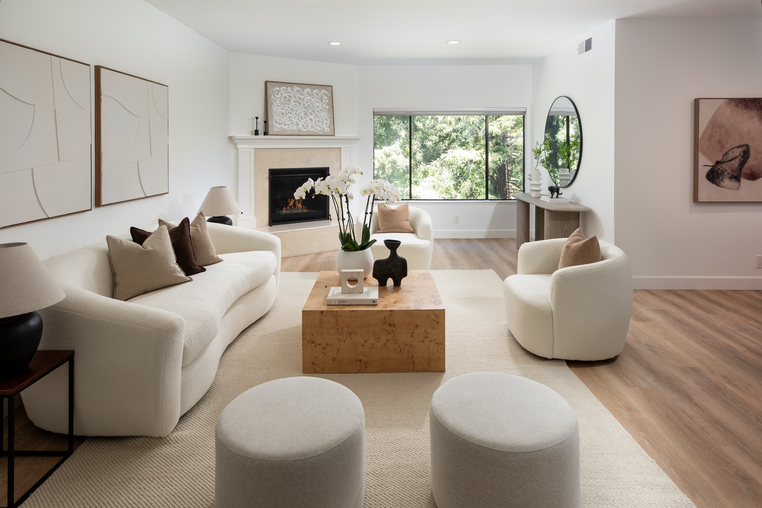 A modern living room with white sofas, a wooden coffee table, a fireplace, large window with greenery outside, wall art, and decorative objects on a console table.