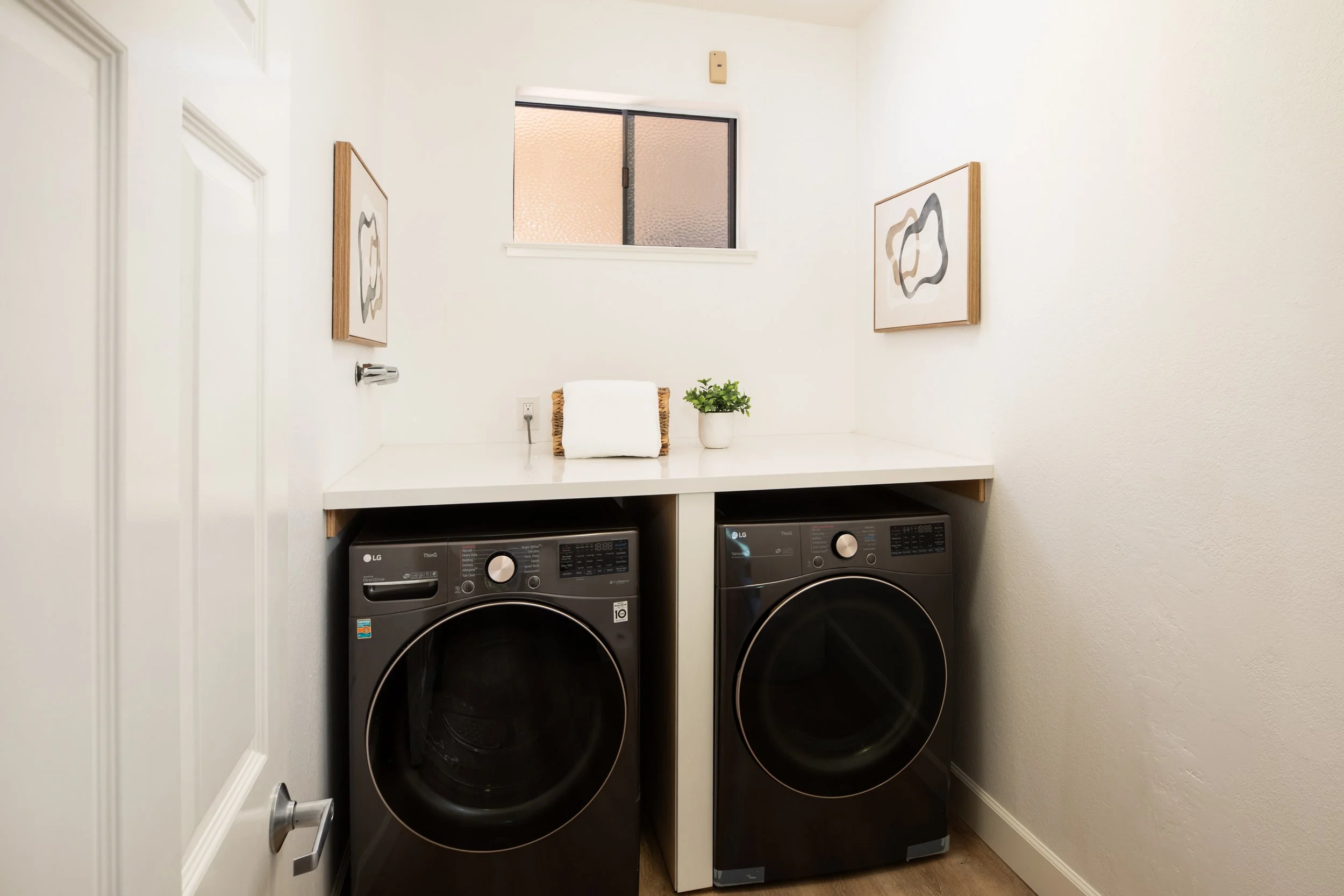 A laundry room with a white countertop, a window, two black front-loading laundry machines, a small plant, and wall art