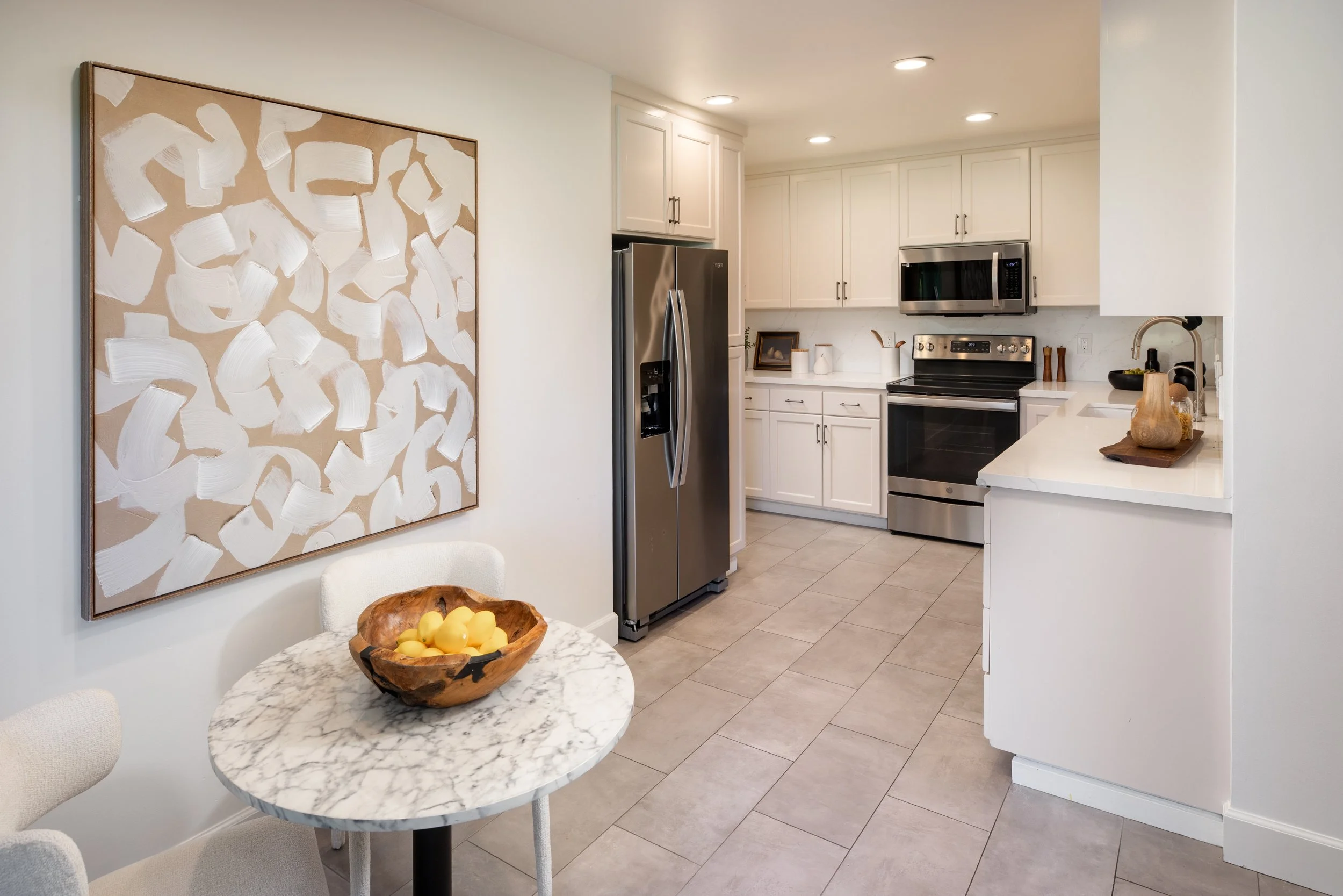 A modern kitchen with white cabinets, stainless steel appliances including a refrigerator, microwave, and oven. There is a framed abstract artwork on the wall, and a small marble-topped dining table with a wooden bowl of lemons on it.