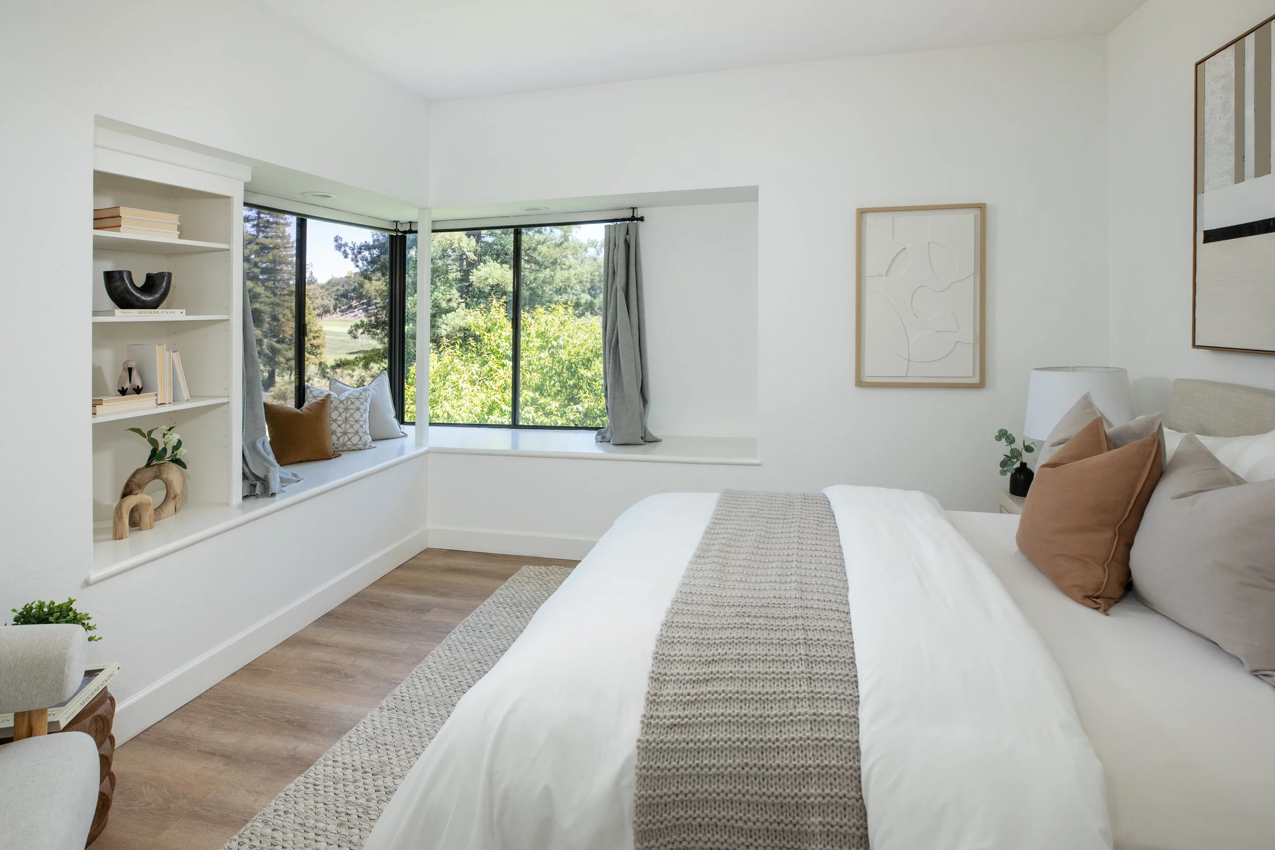 A bright and modern bedroom with large window seating area overlooking greenery, white walls, minimalist artwork, bed with beige and white linens, and a side table with a lamp and small plant.