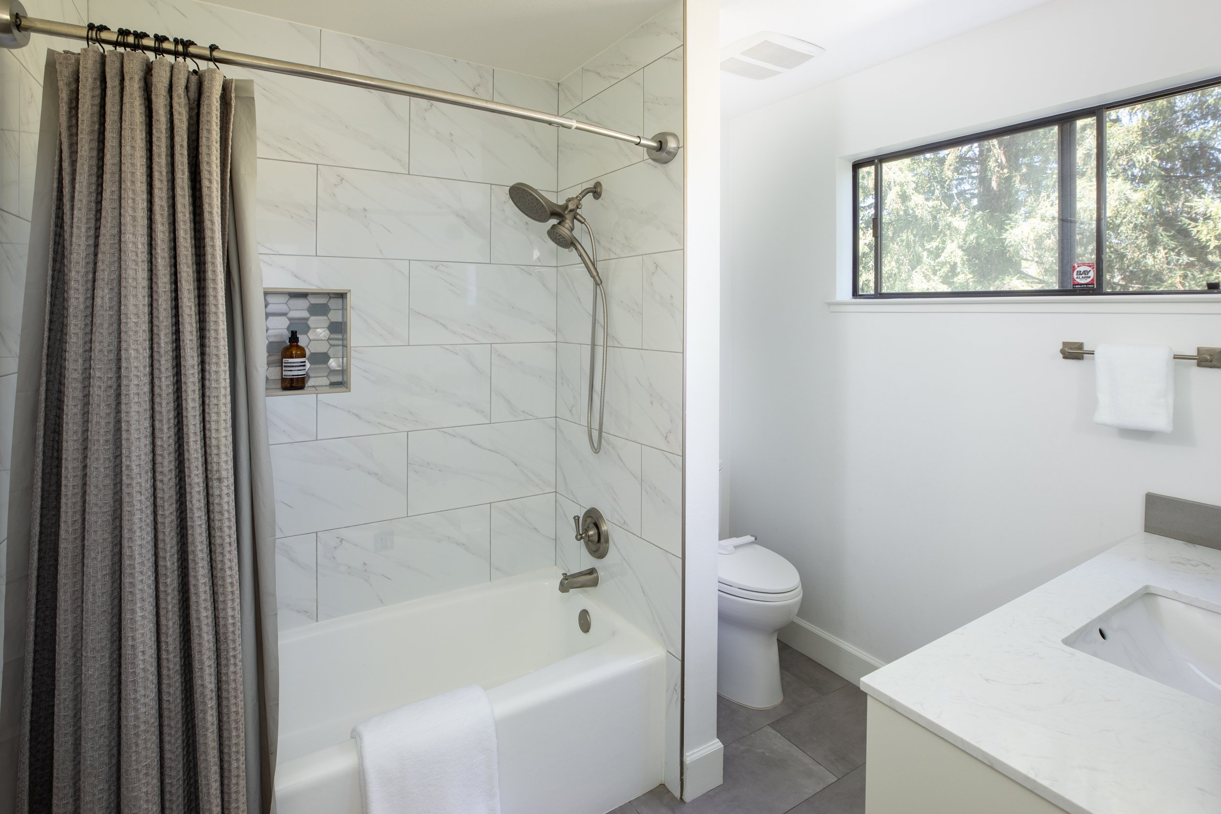 A bathroom with a shower area enclosed with a curtain, marble wall tiles, a built-in soap niche, a showerhead, and a white toilet next to a vanity with a marble countertop and a large window showing outdoor trees.