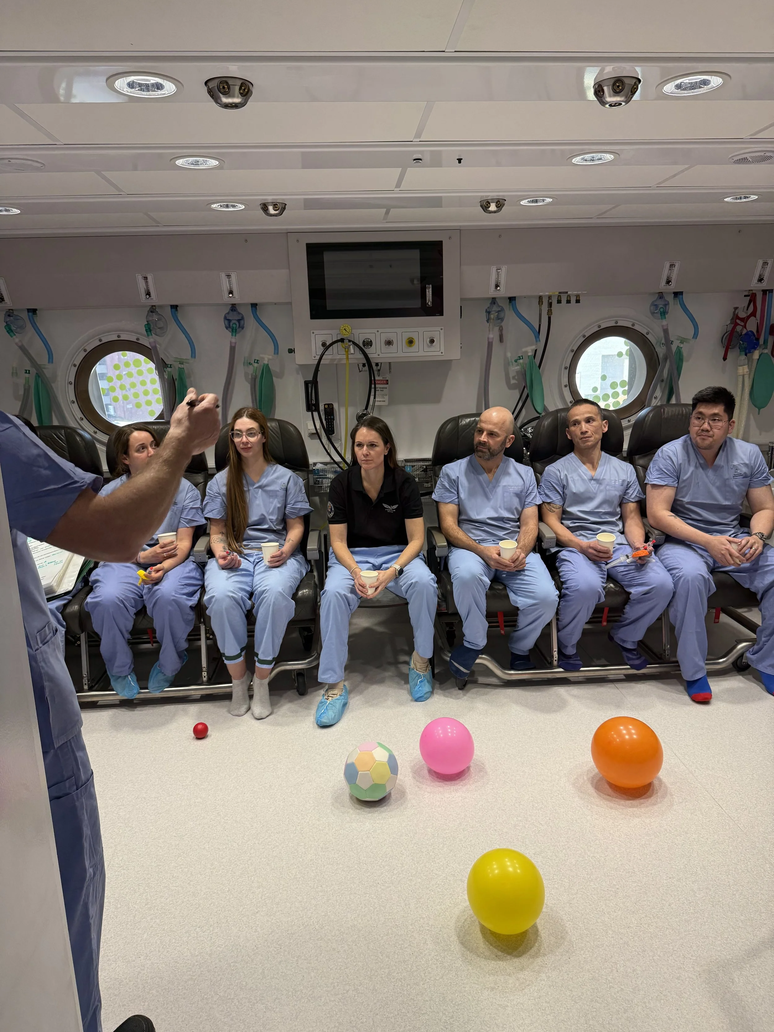 Medical team in blue scrubs seated inside a hyperbaric chamber with colorful balls on the floor