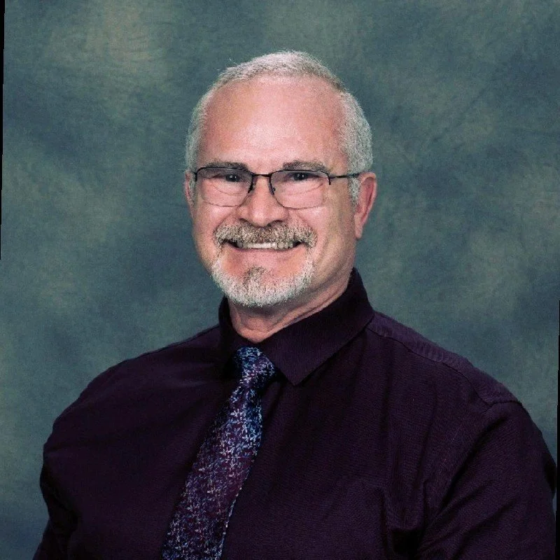 Portrait of a smiling older man wearing glasses, a dark dress shirt, and patterned tie against a blue-gray background.