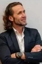 Smiling man with long brown hair wearing a dark blazer and white shirt