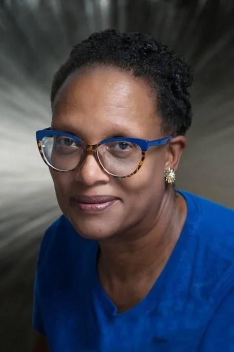 Portrait of a smiling Black woman wearing blue tortoiseshell glasses, floral earrings, and a blue shirt
