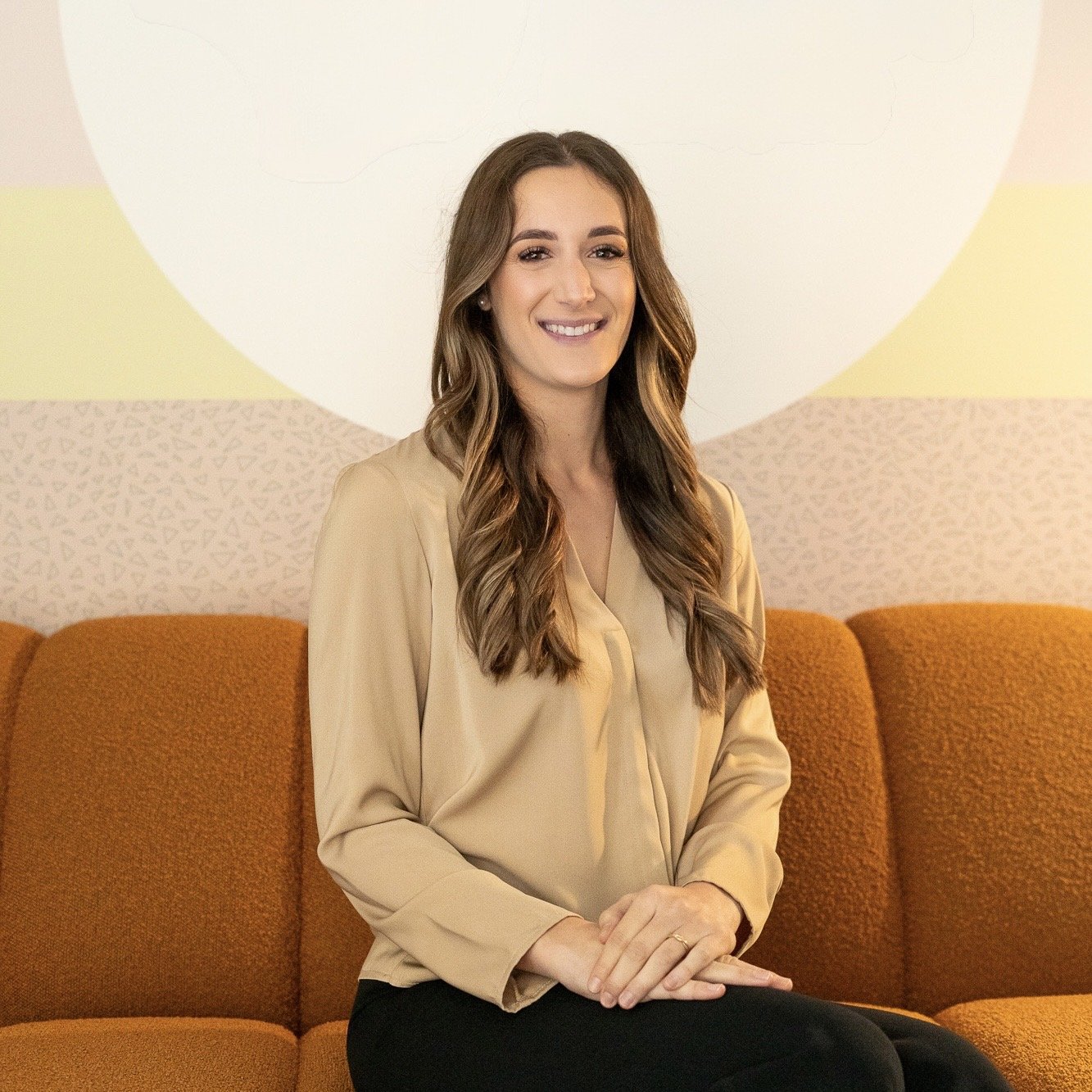 Smiling woman with long brown hair sitting on an orange sofa in a beige blouse