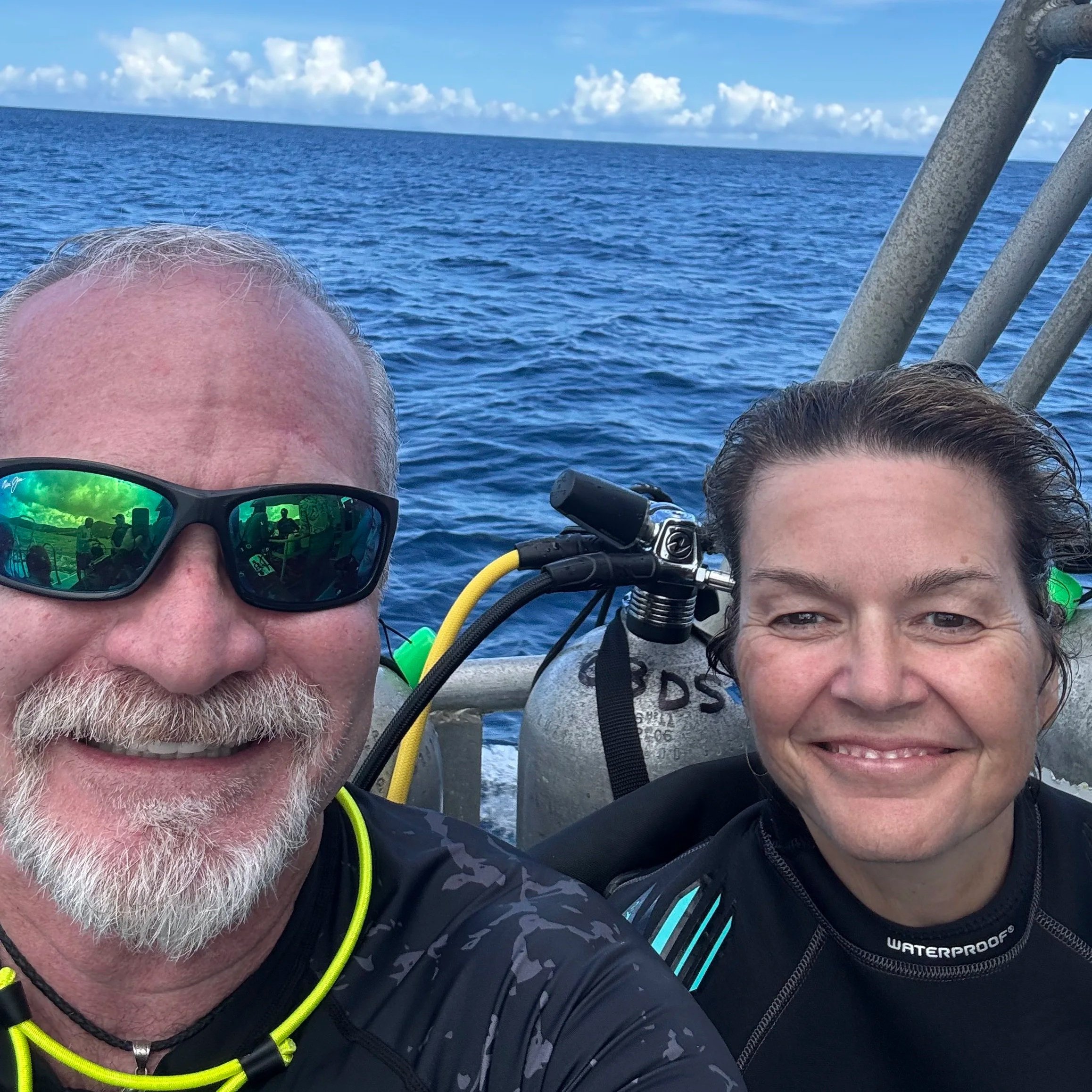 Two scuba divers smile for a selfie on a boat, with scuba gear, blue ocean, and cloudy sky behind them.