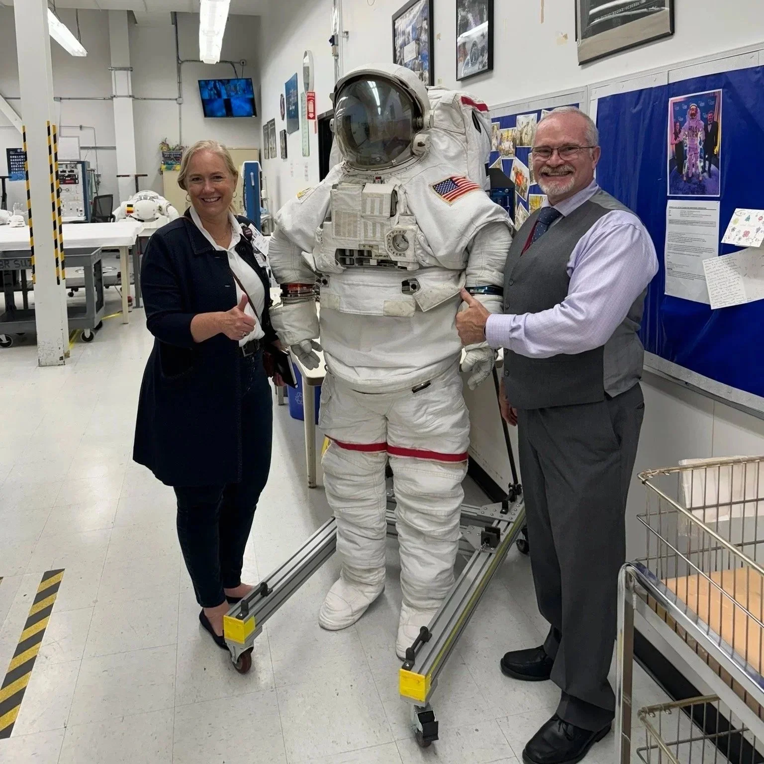 Two people pose beside a white astronaut spacesuit in a space training facility.