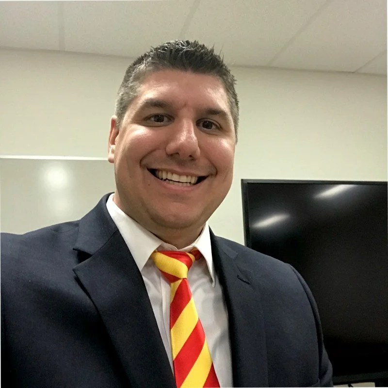Smiling man in a dark suit and red-and-yellow striped tie taking a selfie indoors