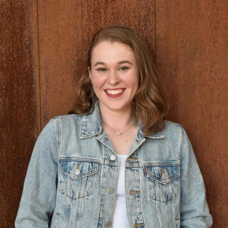 Smiling woman wearing a light denim jacket and white shirt in front of a wood-paneled wall
