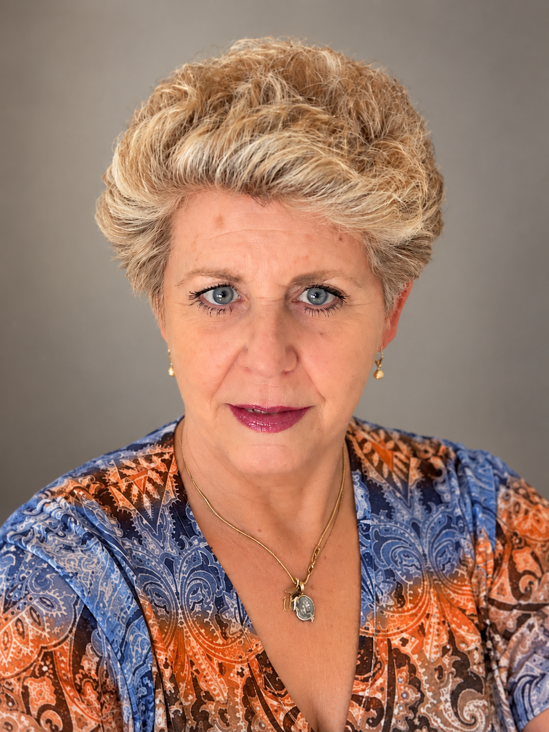 Portrait of an older woman with short blond hair, blue eyes, gold jewelry, and a colorful patterned blouse