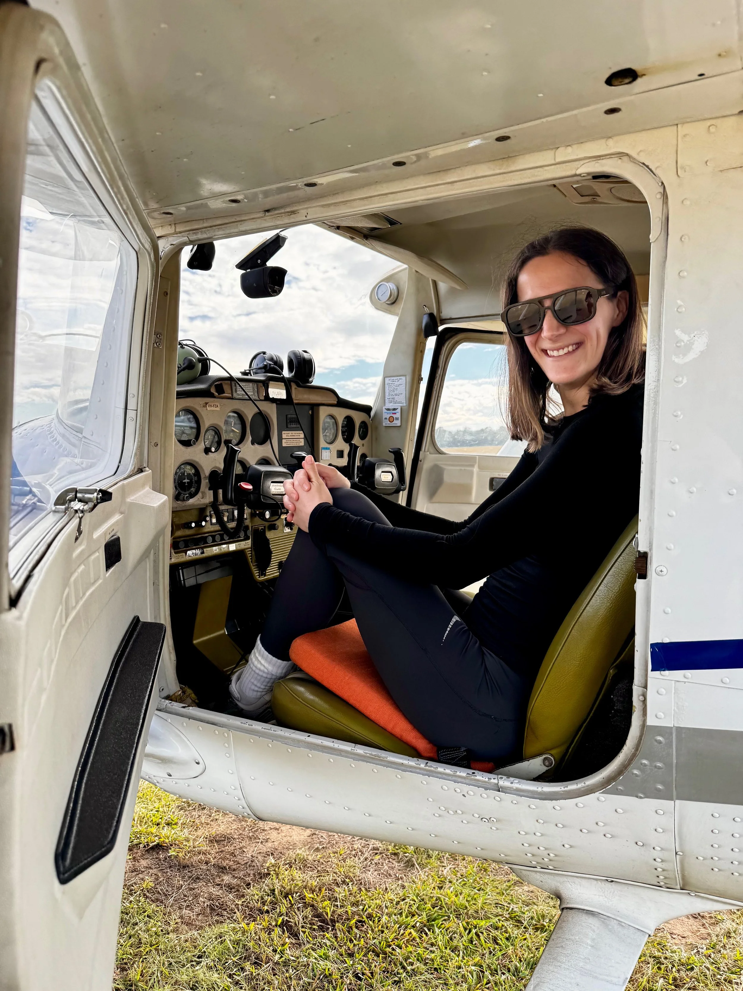 Woman wearing sunglasses sits in the cockpit of a small airplane, with the instrument panel and open door visible.