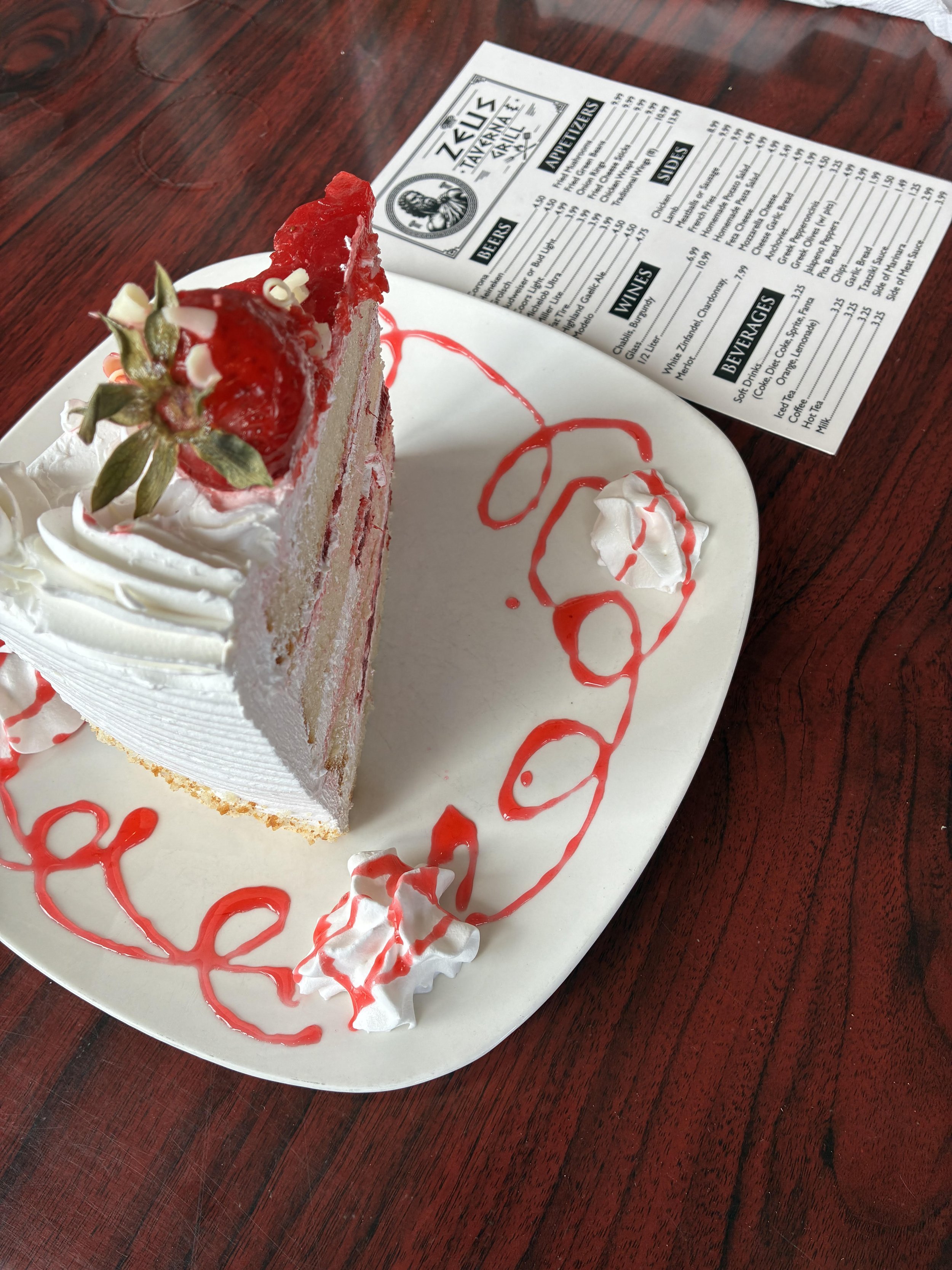 A slice of cheesecake with whipped cream, a strawberry on top, and red sauce drizzled on the plate, on a white plate with a red wood table in the background.