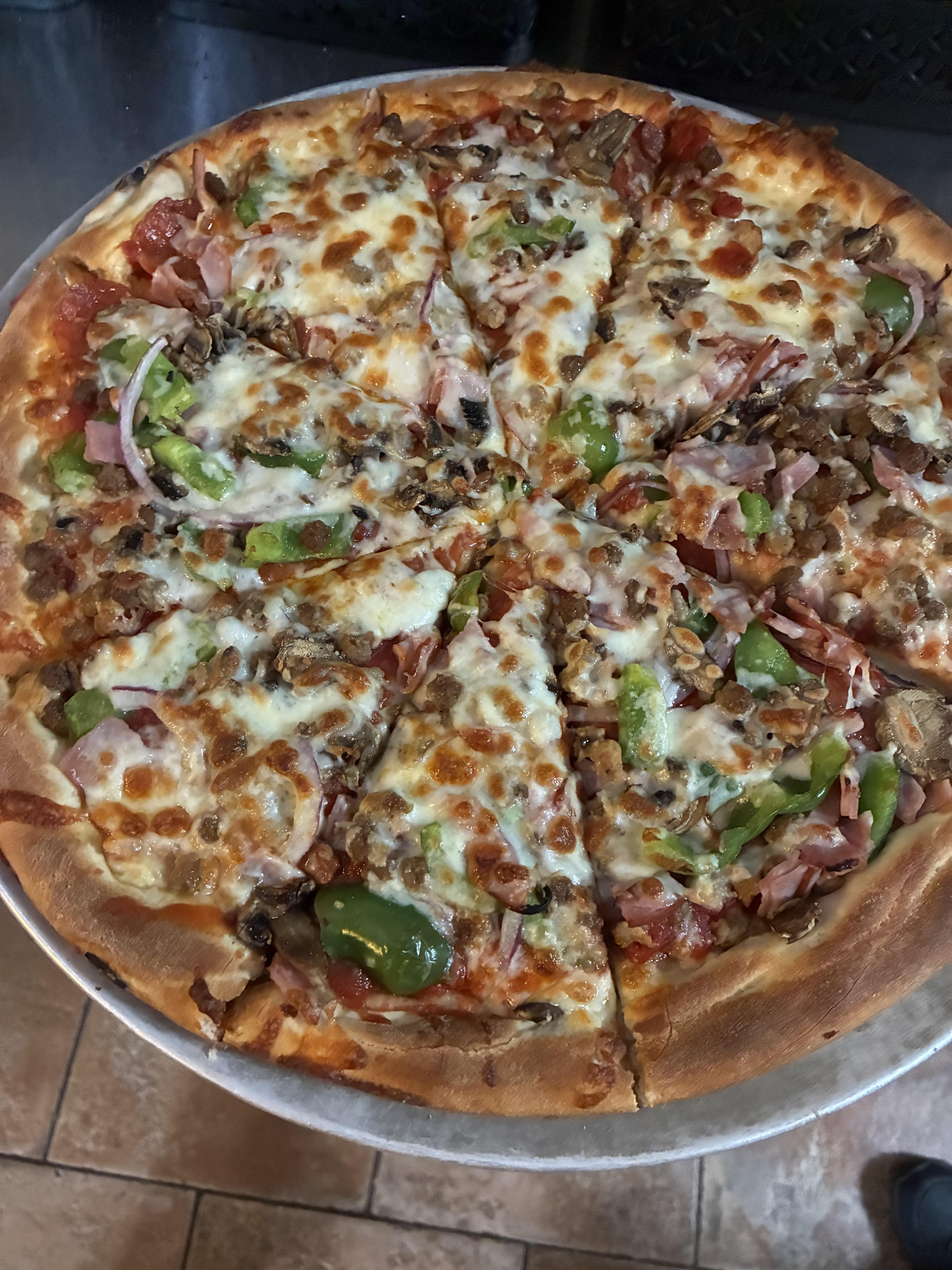 A large pizza on a metal tray topped with cheese, green peppers, red onions, mushrooms, pepperoni, ham, sausage, and bacon.