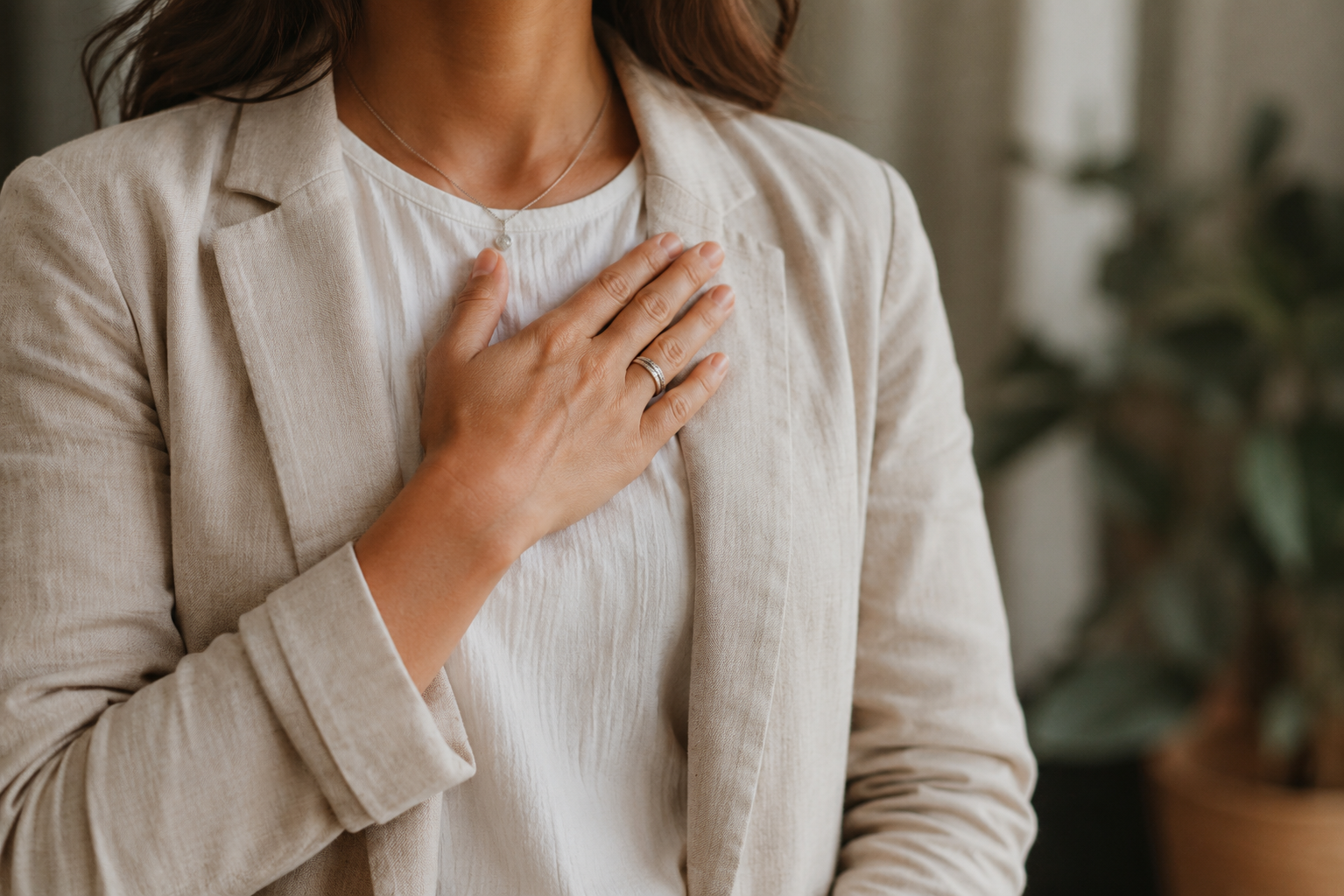 A woman with light skin and brown hair, wearing a beige blazer and a white shirt, has her left hand on her chest, wearing a wedding ring and a necklace.