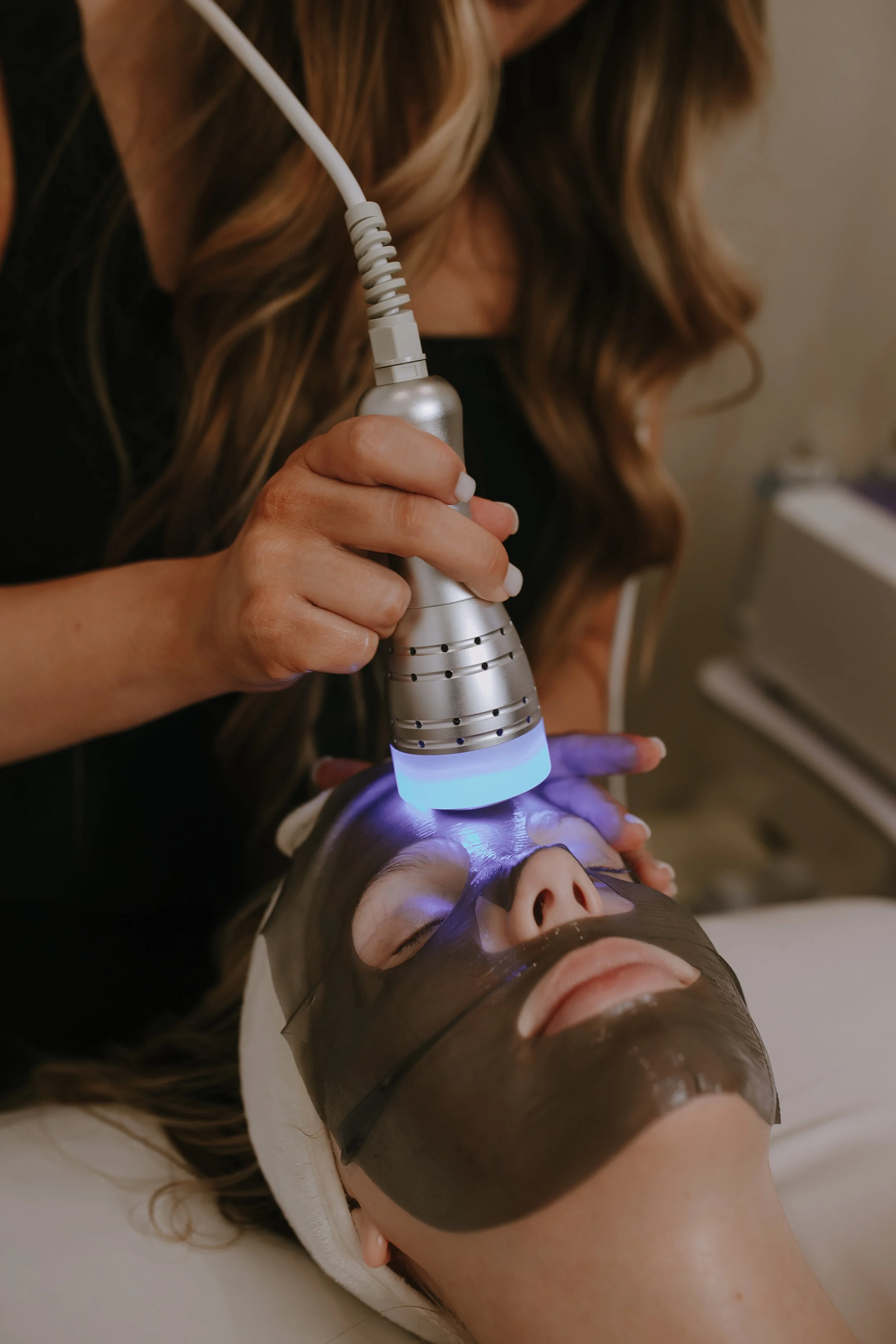 A woman performing a facial treatment using a handheld device emitting blue light on a patient's face, who is lying down with eyes closed and protective eye mask, in a clinical setting.
