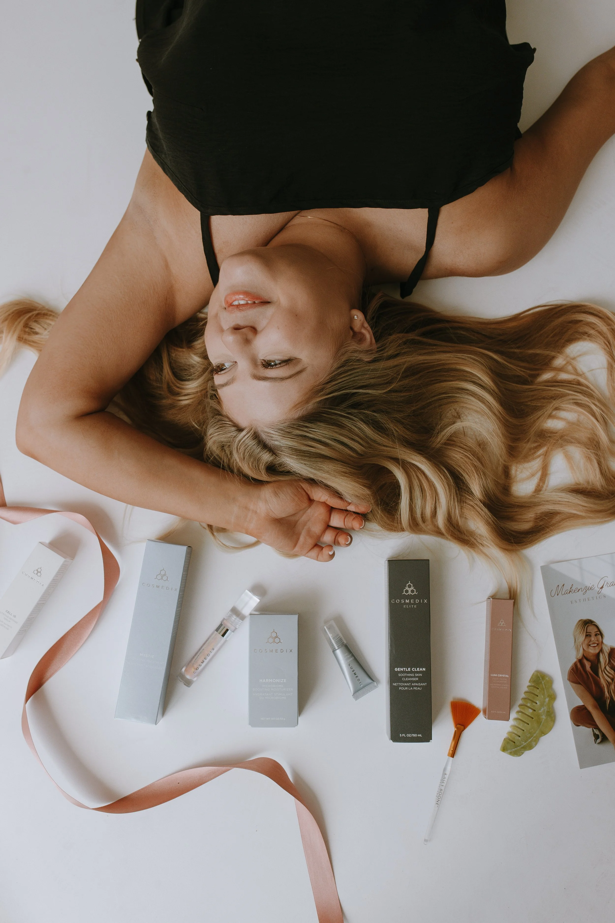 A woman with long blonde hair lying on a white surface, wearing a black top. Various skincare or cosmetic products are arranged around her, including tubes, boxes, and small bottles, some with spray applicators and labels, along with some decorative items like a ribbon, a small orange spatula, and green leaf-shaped accessories.