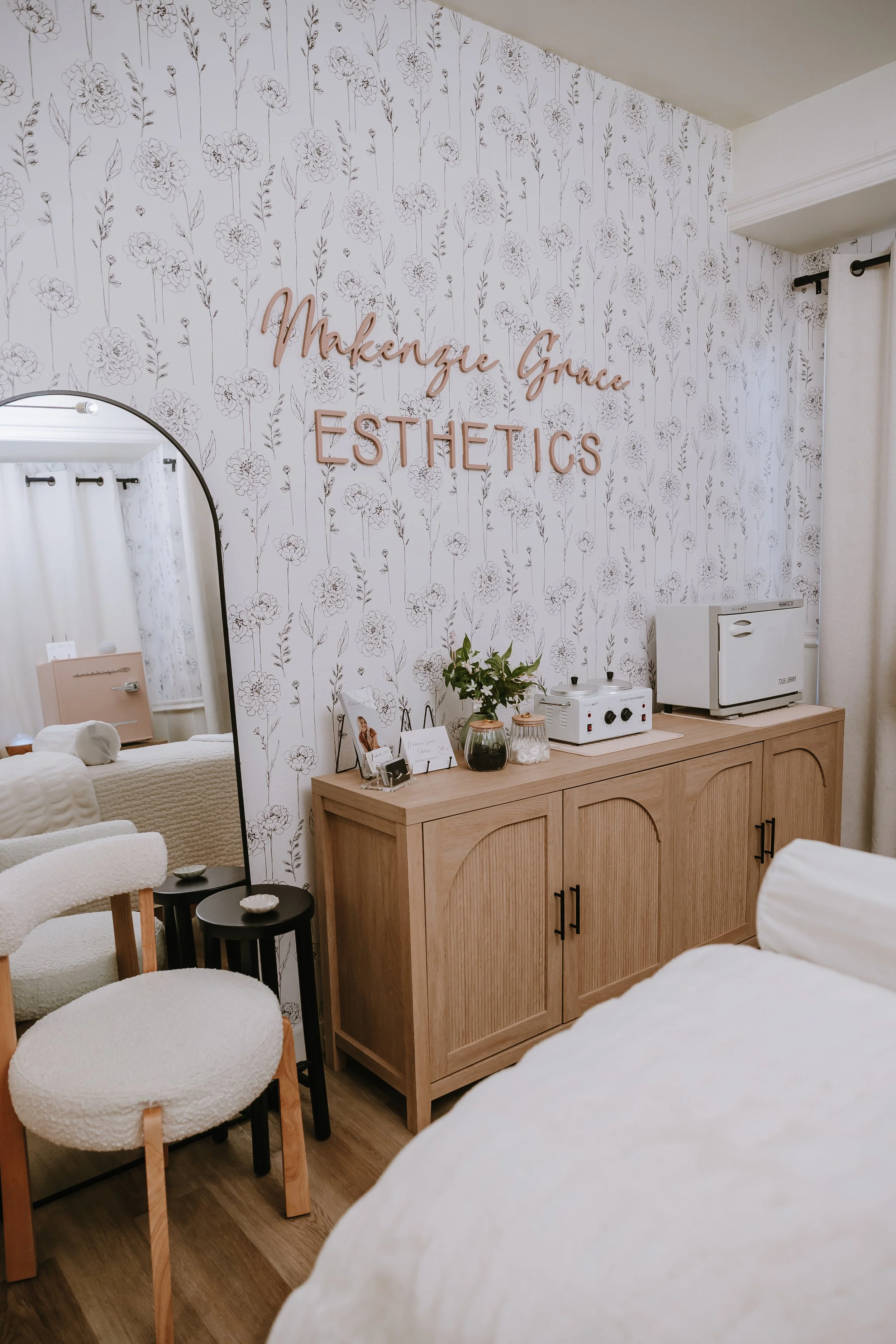 Interior of a spa or aesthetics clinic with a wall featuring floral wallpaper, a pink sign that reads 'Makenzi Grove AESTHETICS,' a wooden cabinet with skincare or treatment equipment, and a mirror reflecting a treatment room with a bed and nightstand.