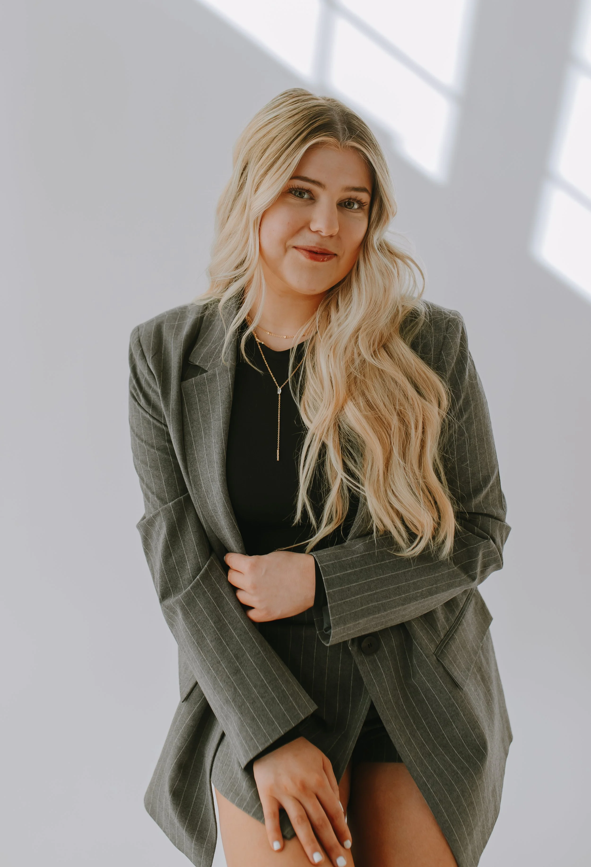 A woman with long blonde hair and green eyes in a gray pinstripe suit, holding her skirt, revealing her thigh and part of her leg, with a neutral expression against a plain background with light coming through windows.